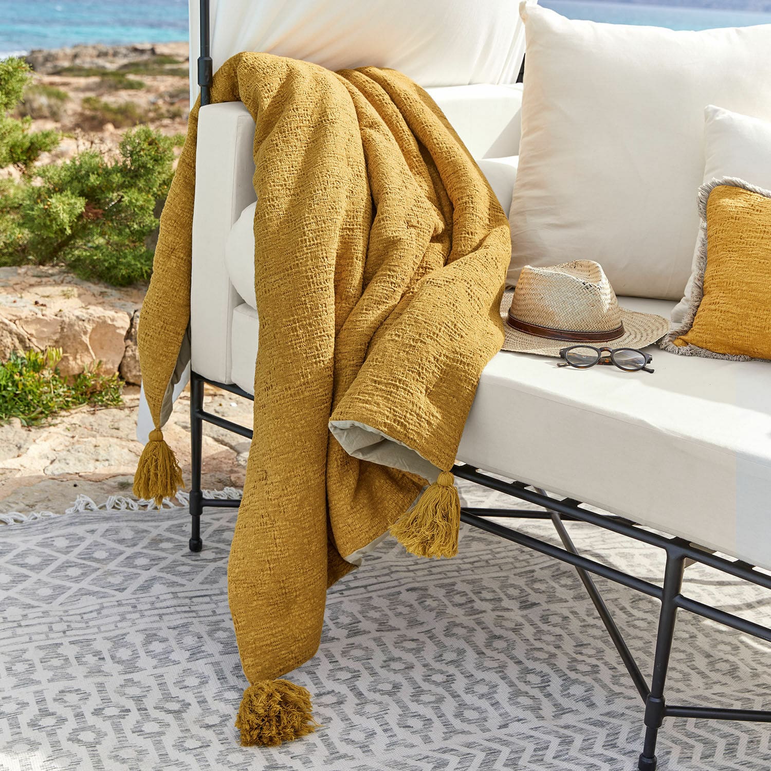 Plaid Rabea Plaid jaune moutarde à pompons sur canapé blanc extérieur, ambiance terrasse avec lunettes et chapeau