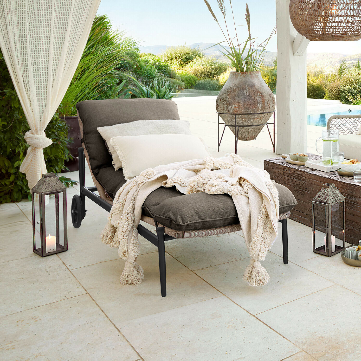 Chaise longue de jardin en tissu gris foncé avec couverture crème à franges sur terrasse avec vue sur jardin