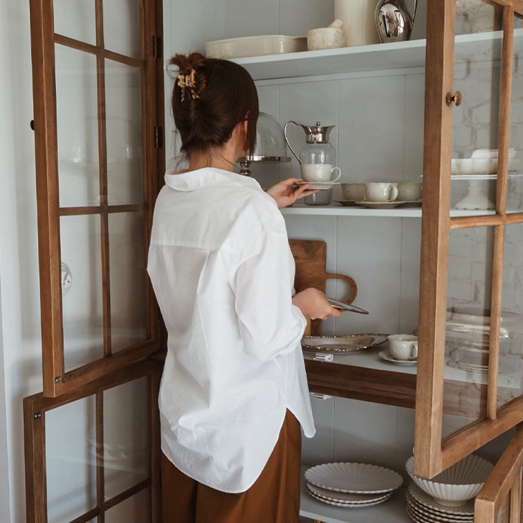 Femme en chemise blanche prenant une tasse en porcelaine dans une vitrine en bois avec vaisselle