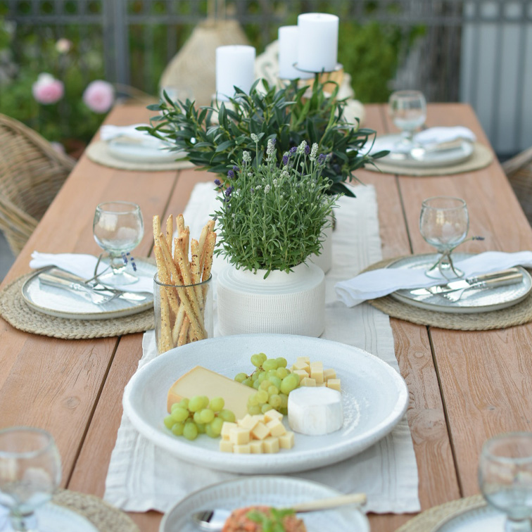 Table en bois avec vaisselle blanche, fromages, raisins verts et décoration végétale pour repas en plein air