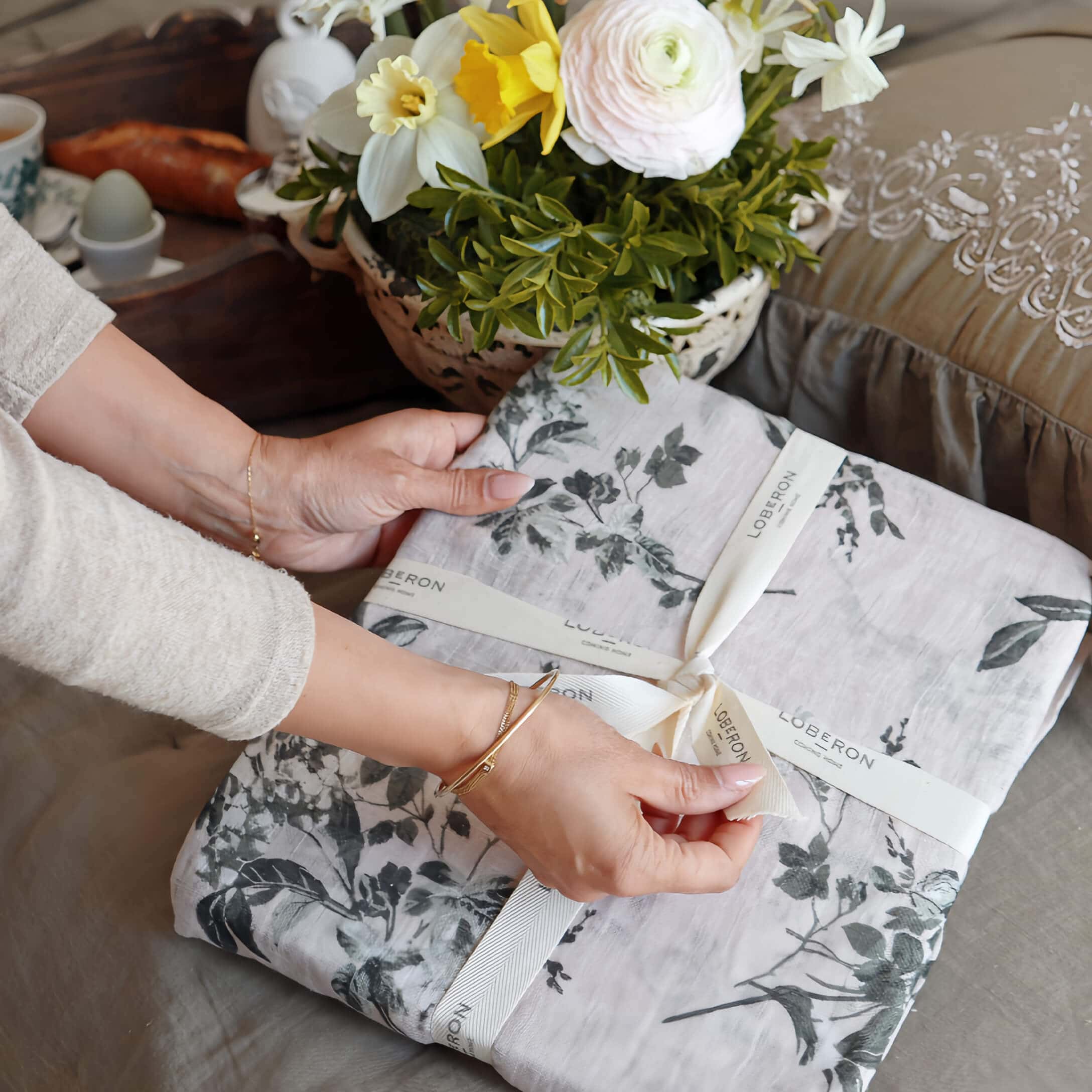 Mains déballant un linge de lit en lin gris à motif végétal, bouquet de fleurs en arrière-plan