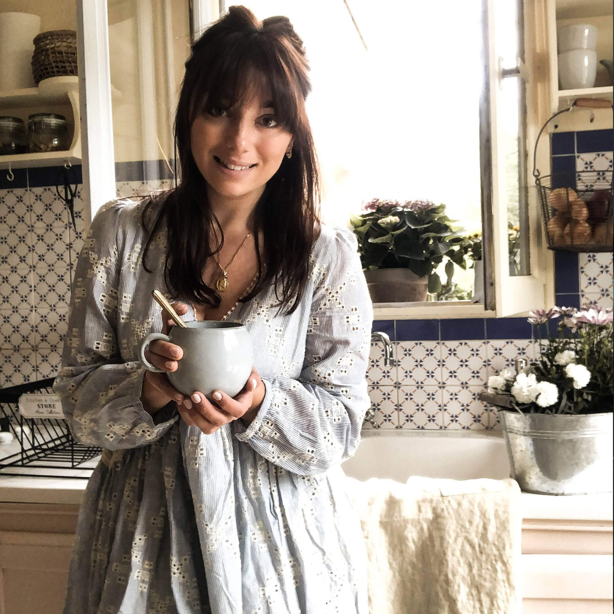 Femme en robe claire tenant une tasse en céramique dans une cuisine avec carrelage bleu et blanc et fleurs