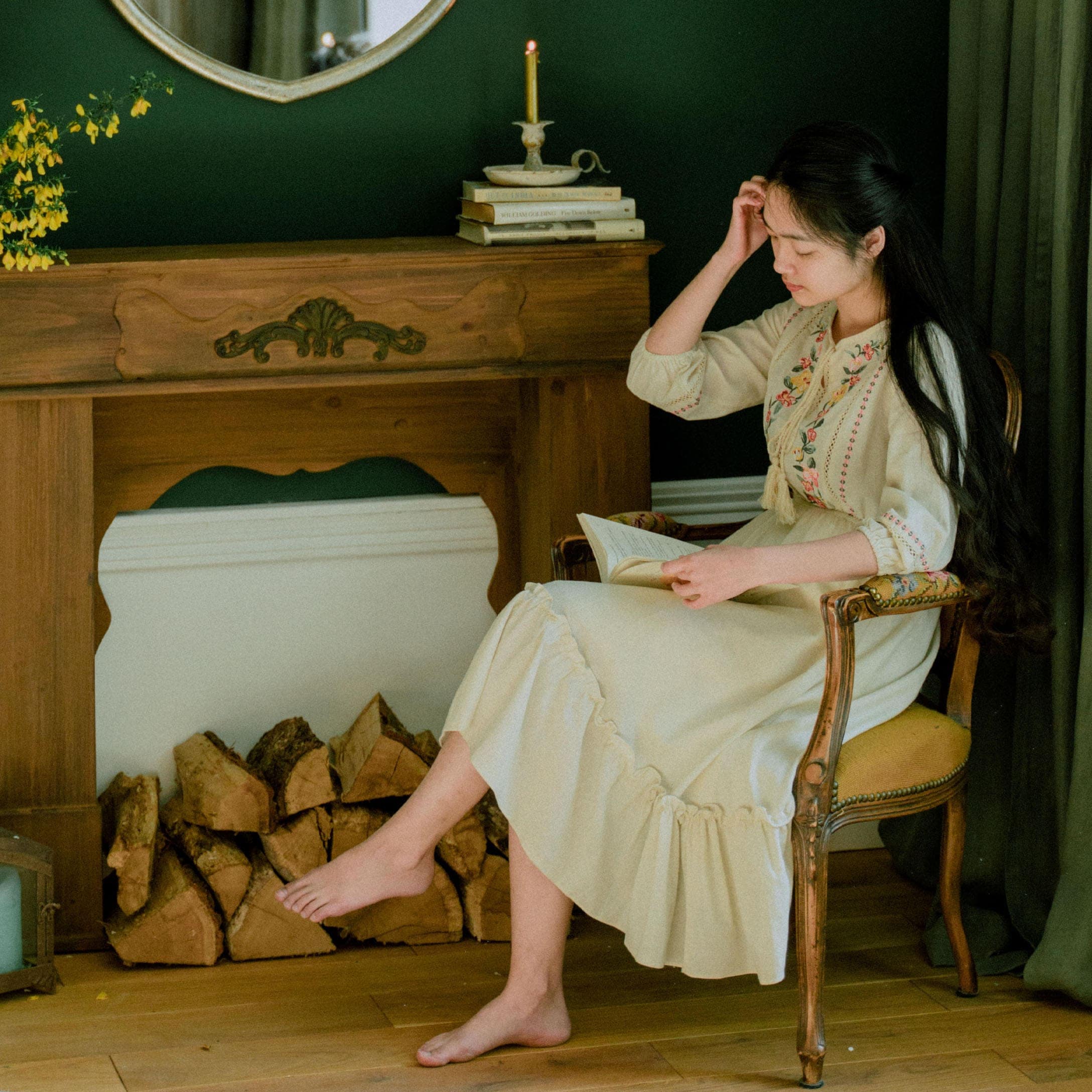 Femme en robe crème brodée assise sur une chaise en bois près d'une cheminée avec du bois