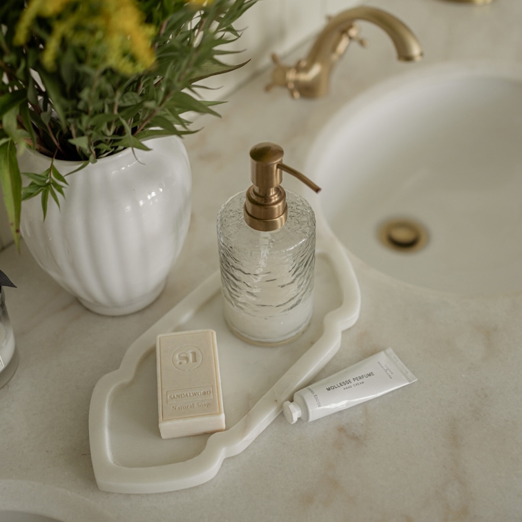 Distributeur de savon en verre avec pompe en laiton sur plateau blanc avec savon et crème pour les mains sur lavabo en marbre