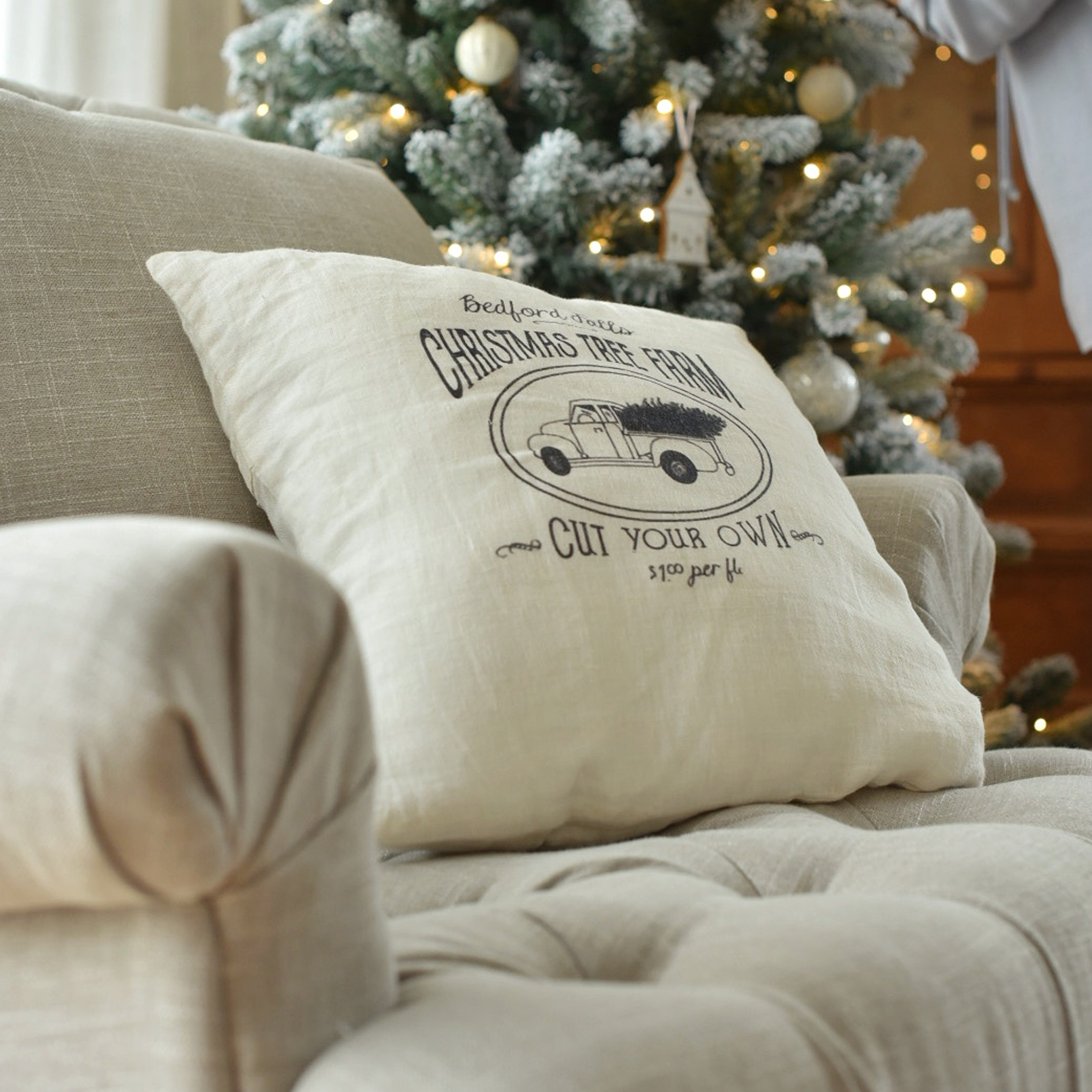 Coussin beige en lin avec imprimé camion de Noël sur canapé près d'un sapin décoré de lumières