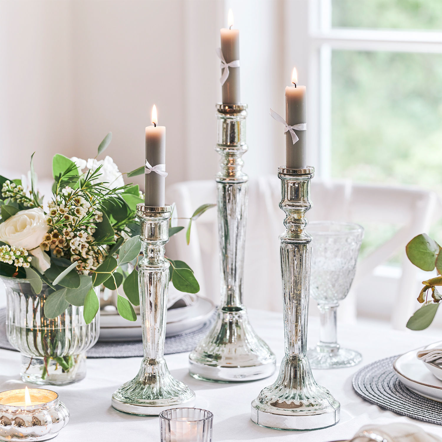 Lot de 3 bougeoirs Riley Trois chandeliers argentés avec bougies grises sur une table blanche décorée de fleurs et de verre