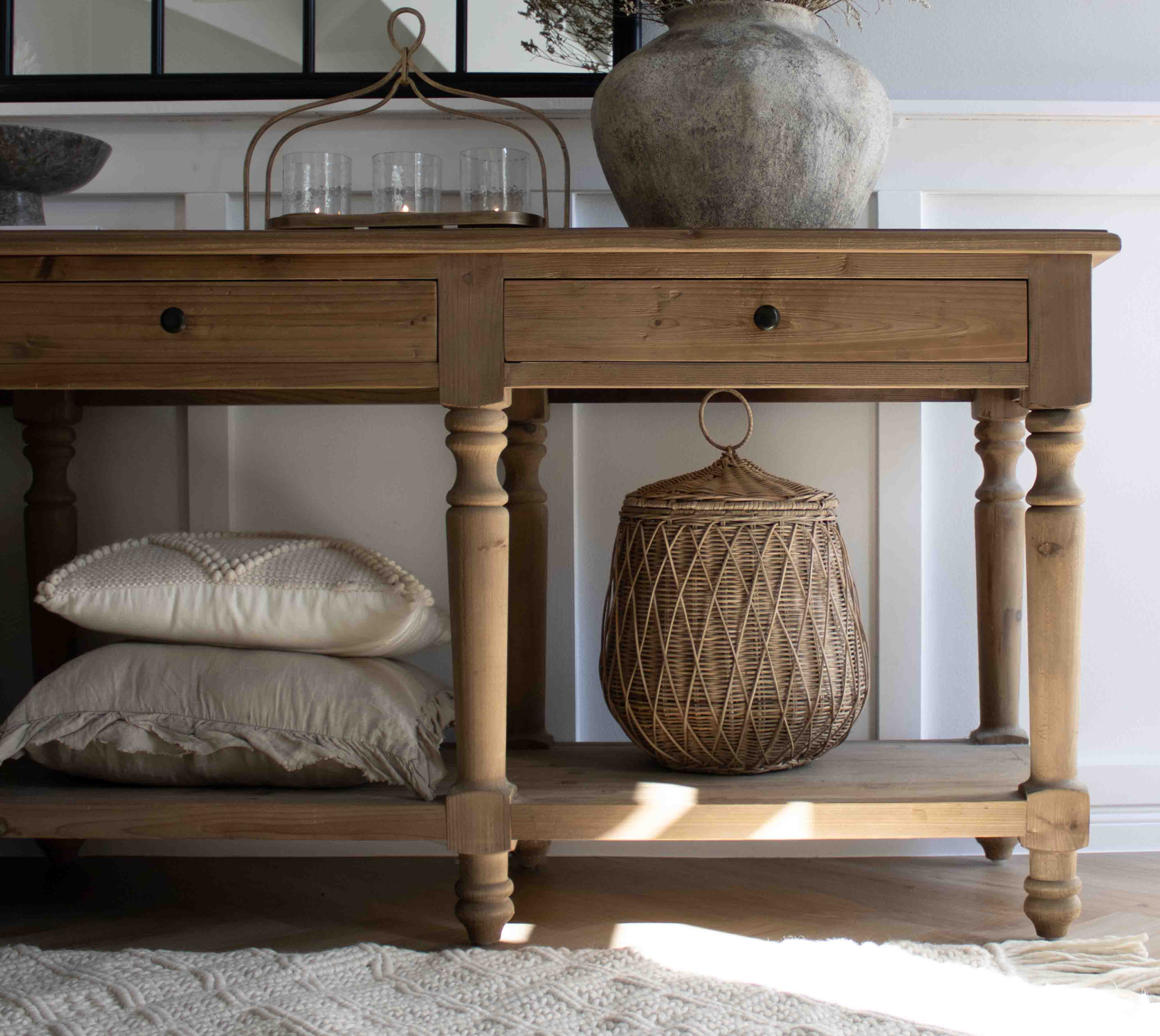 Table en bois naturel avec tiroirs, panier en osier et coussins empilés en dessous dans un intérieur cosy