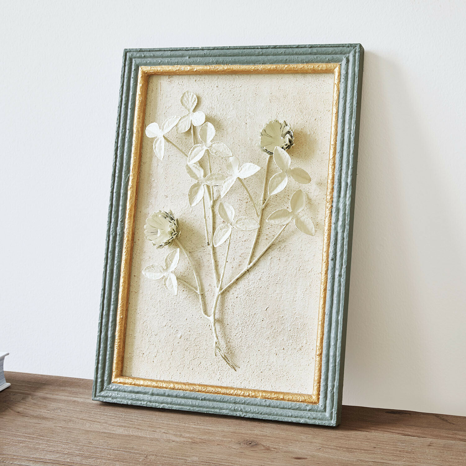 Décoration murale Rivolenne Tableau décoratif avec relief floral dans un cadre vert et doré posé sur une table en bois
