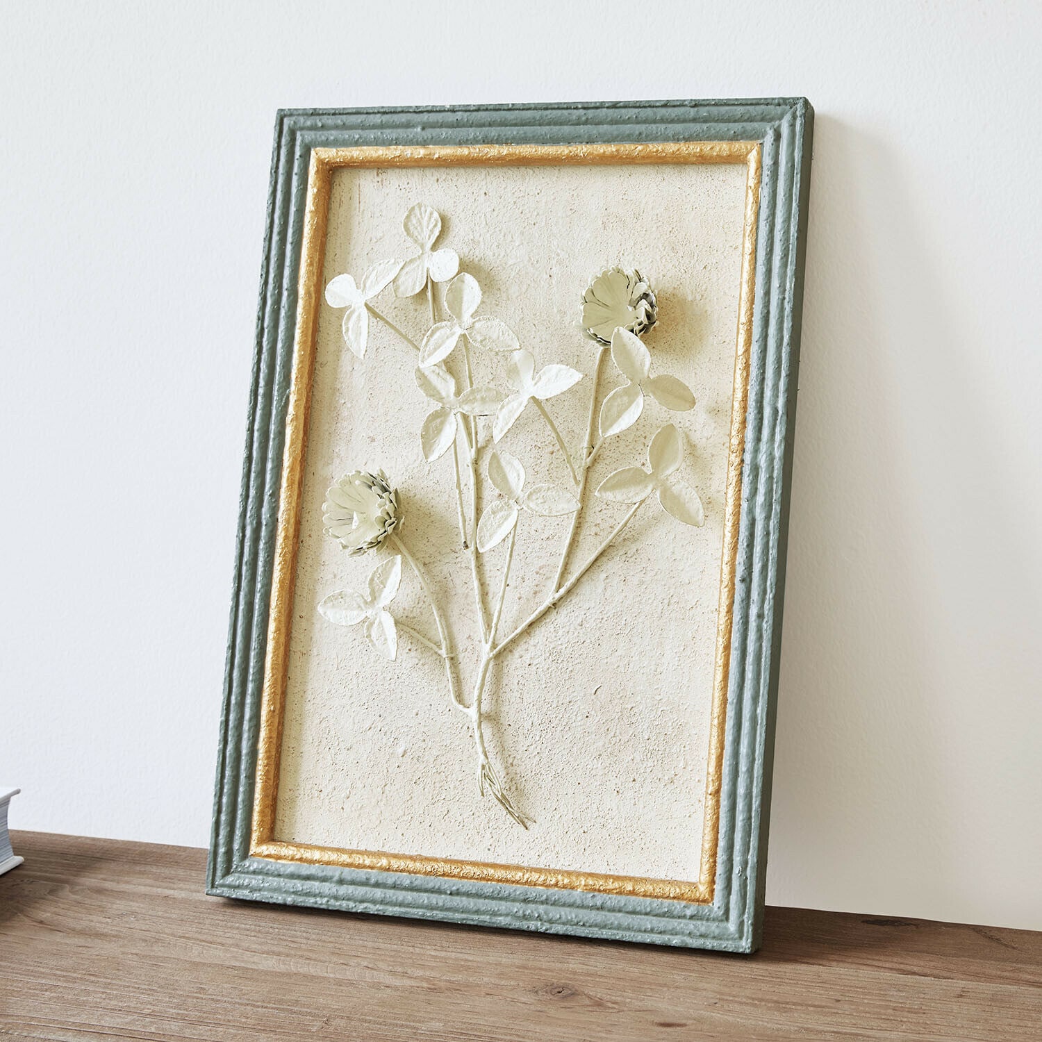 Décoration murale Rivolenne Tableau décoratif avec relief floral dans un cadre vert et doré posé sur une table en bois