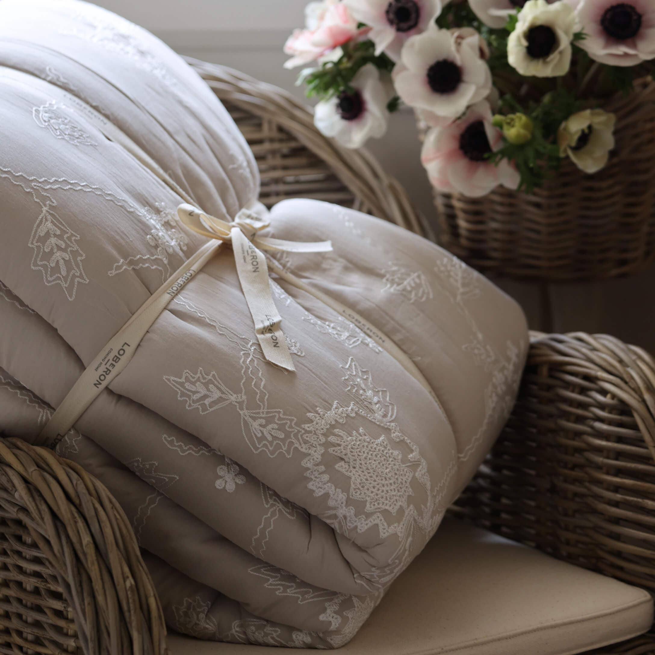Couette beige brodée posée sur un fauteuil en osier avec un panier de fleurs, décoration intérieure cosy