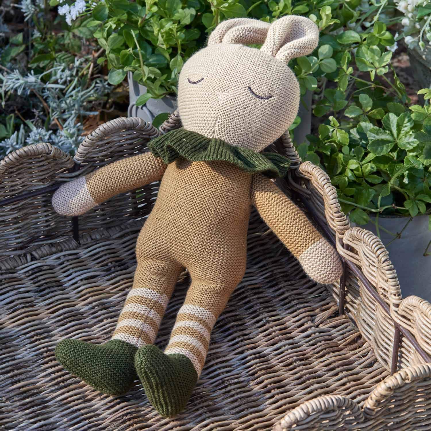 Jouet lapin tricoté beige avec chaussures et col verts sur fond en osier, décoration enfantine