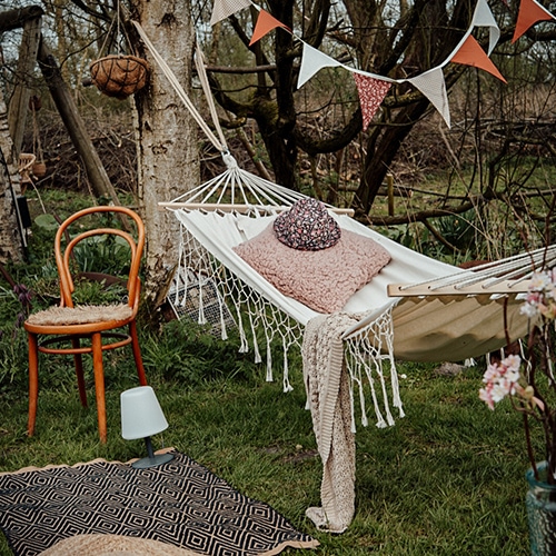 Hamac en tissu blanc avec coussin rose et plaid sur pelouse verte, chaise en bois et guirlande décorative
