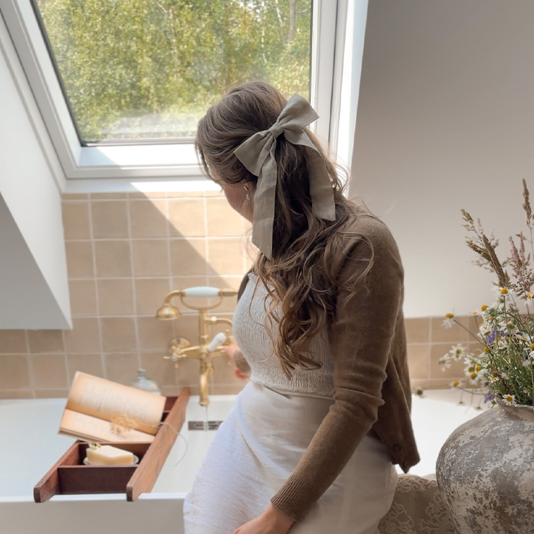 Femme avec un nœud en lin beige dans les cheveux, assise près d'une baignoire avec tablette en bois et livre, lumière naturelle