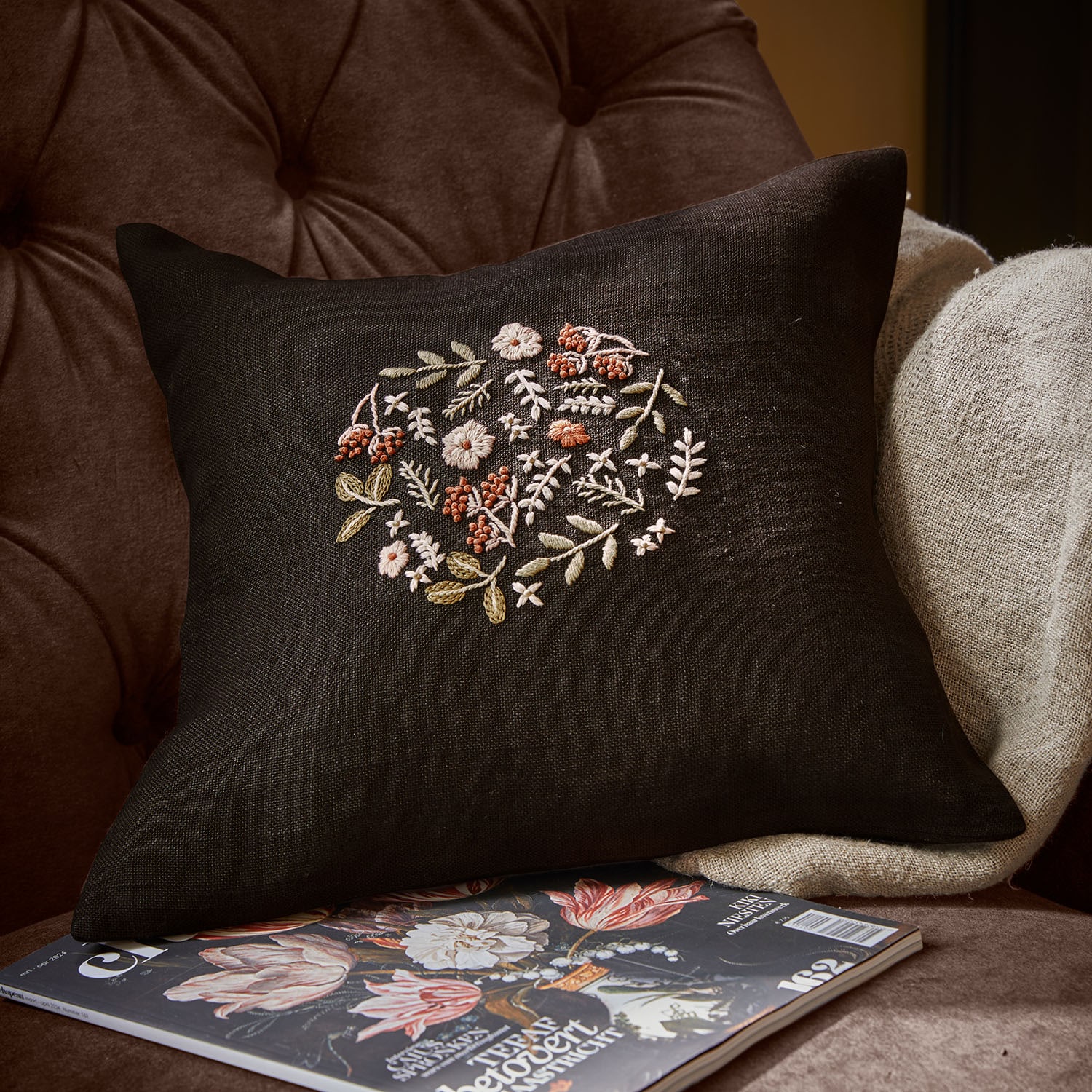 Coussin en lin noir avec broderie florale pastel posé sur un fauteuil marron confortable