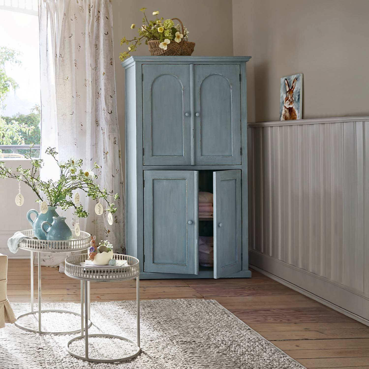 Armoire en bois bleu à deux portes dans un intérieur lumineux avec décorations florales et tapis