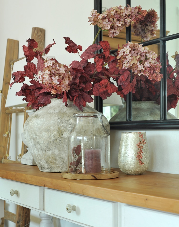 Vase décoratif en pierre avec feuilles rouges et fleurs roses sur table en bois devant un miroir