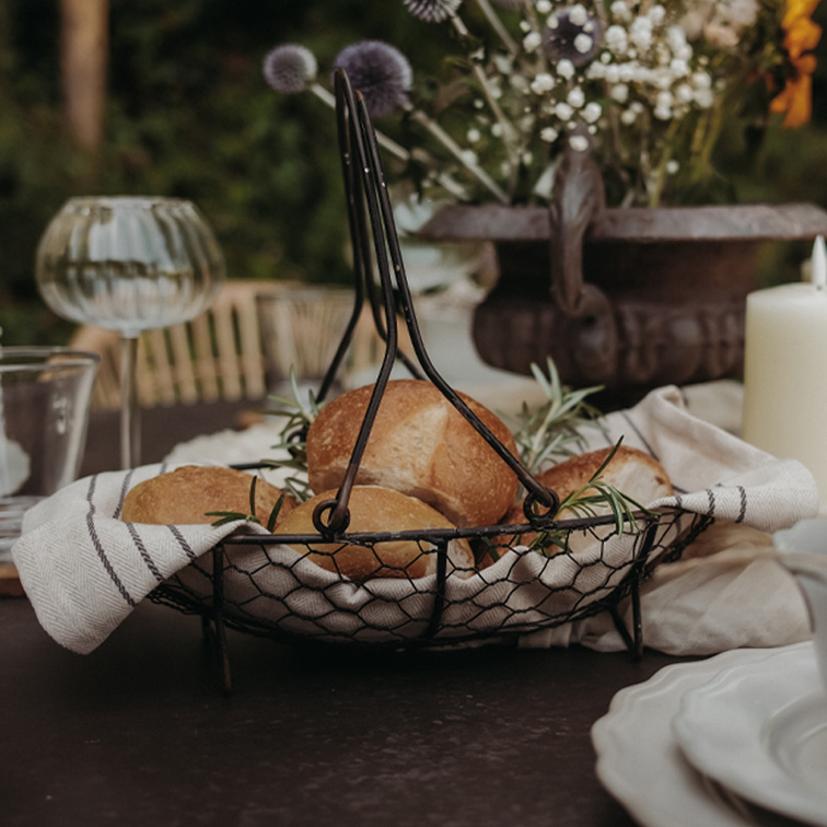 Panier en métal avec poignées, doublé d’un torchon en lin, contenant des petits pains et des herbes sur une table