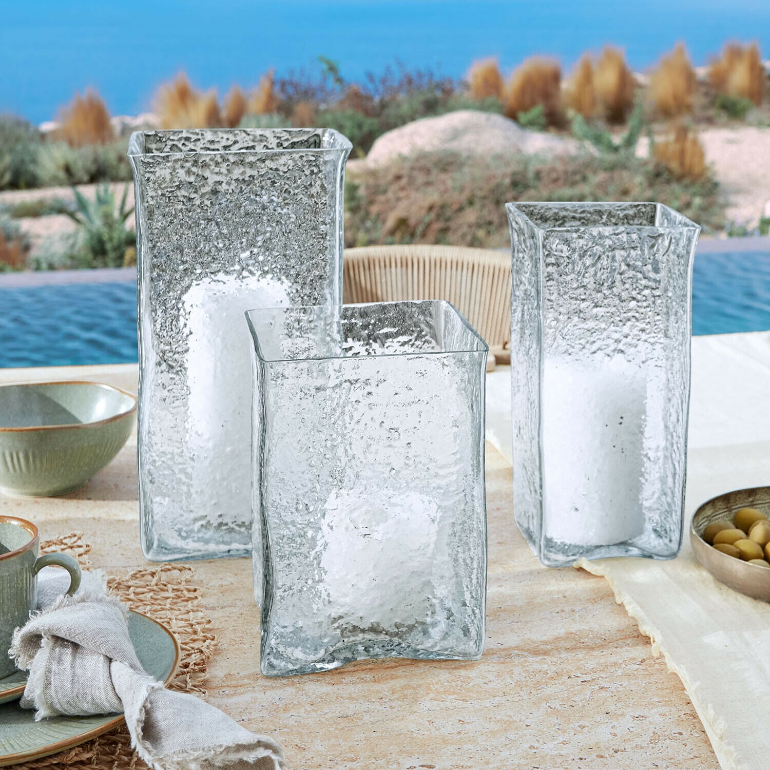 Lot de 3 photophores Xarlac Trois lanternes en verre transparent texturé avec bougies blanches posées à l'extérieur près de la piscine