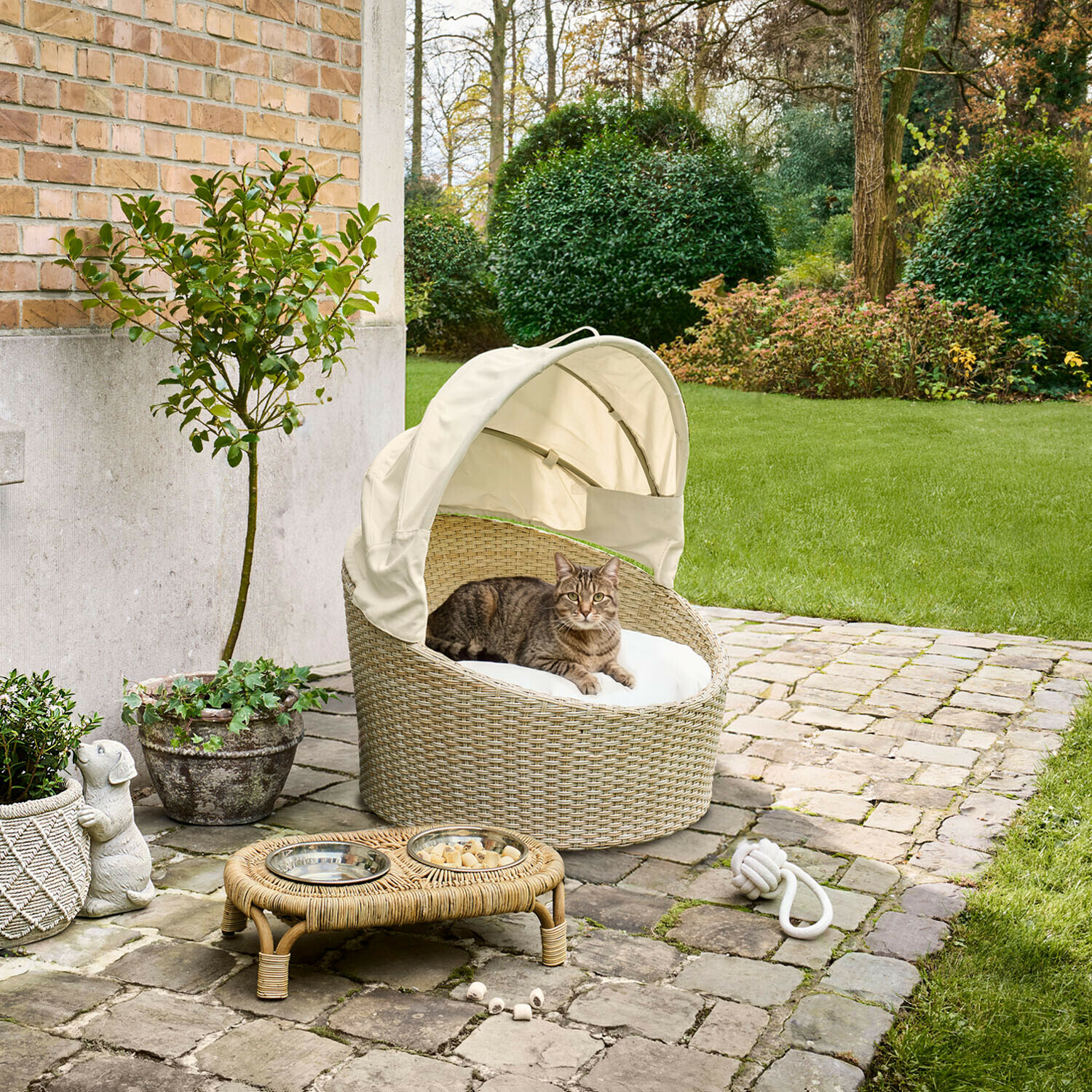 Panier d’intérieur et d’extérieur pour animaux Brinelza Panier ovale en rotin beige avec coussin blanc et auvent pour chat, installé sur une terrasse en pierre