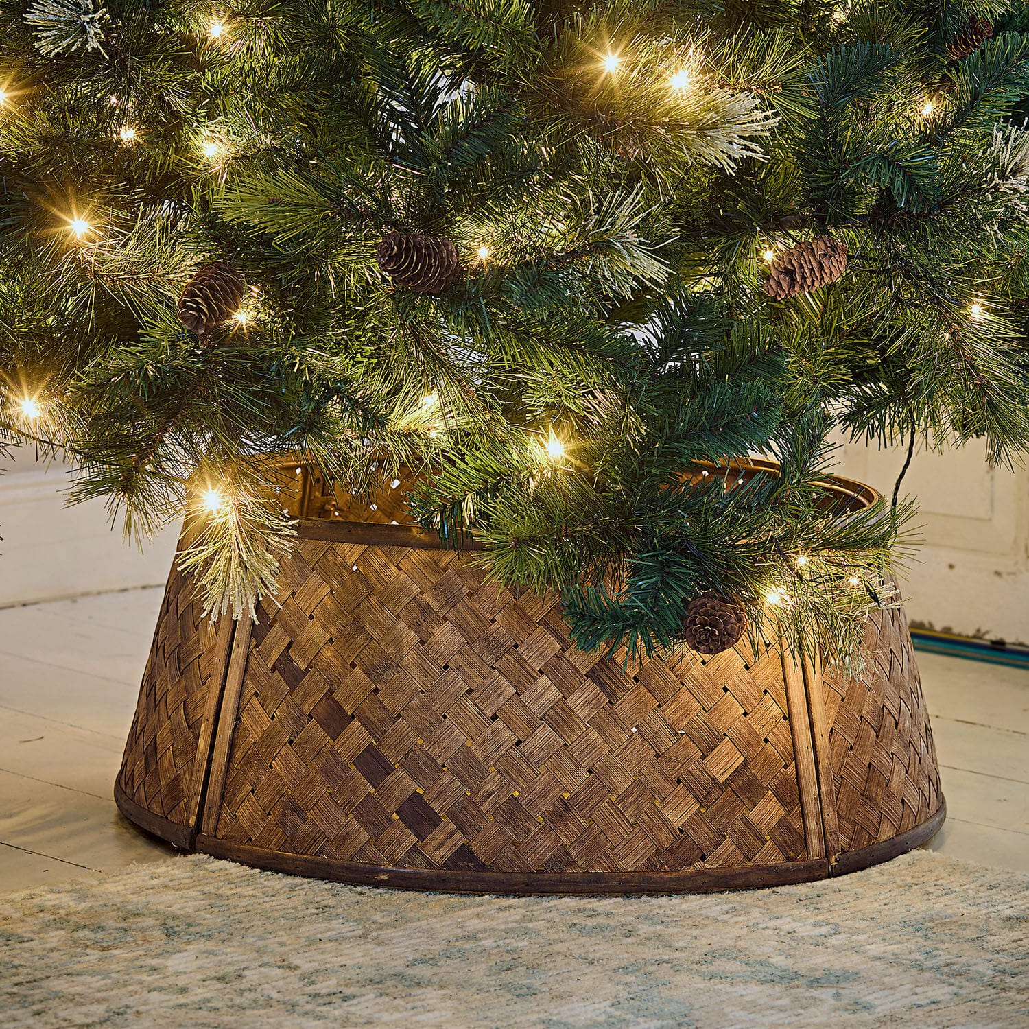 Cache-pied pour sapin de Noël Eden Support de sapin en bois tressé marron sous un sapin vert décoré de lumières et pommes de pin