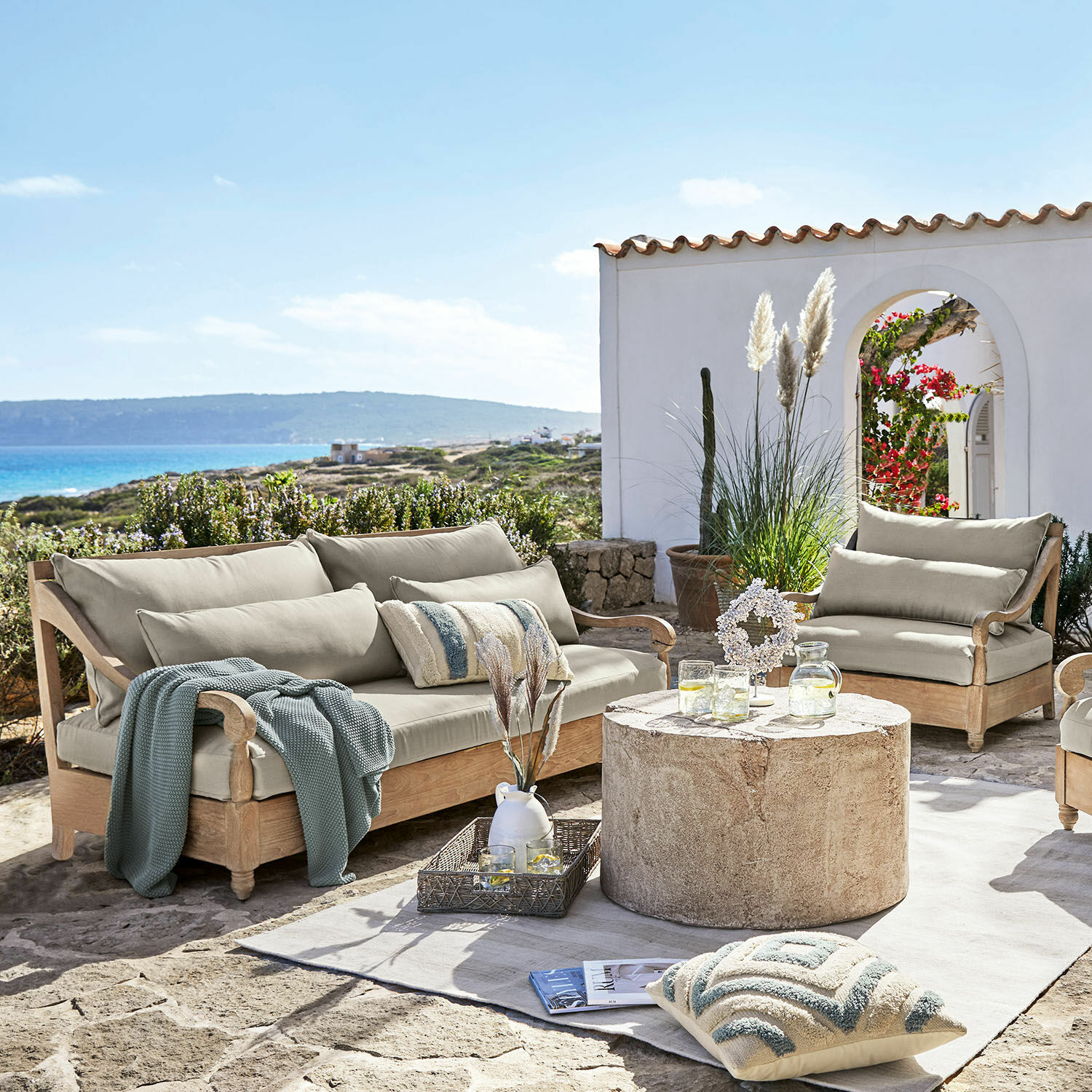 Salon de jardin Montcassin Ensemble de mobilier de jardin en bois clair avec coussins beige sur terrasse en pierre face à la mer