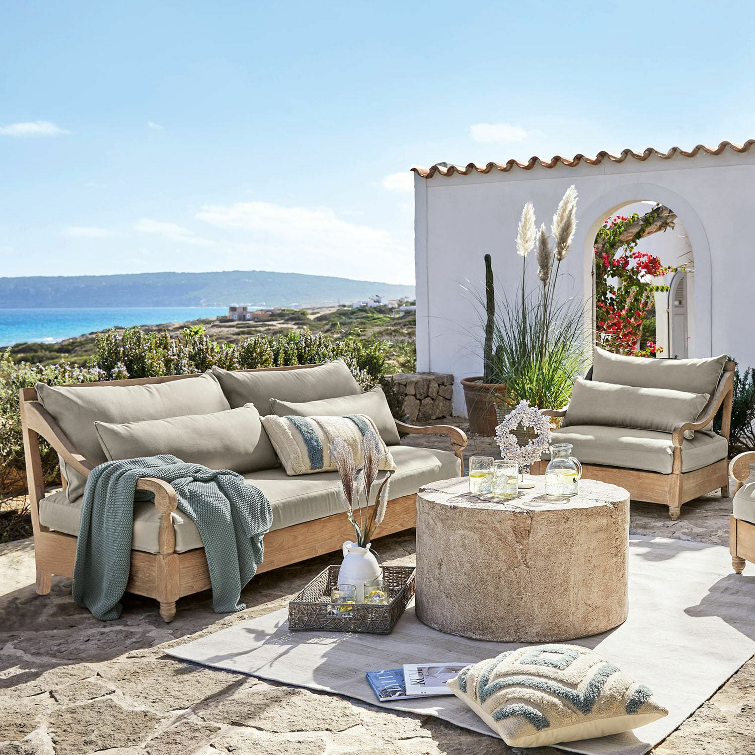 Salon de jardin Montcassin Ensemble de mobilier de jardin en bois clair avec coussins beige sur terrasse en pierre face à la mer