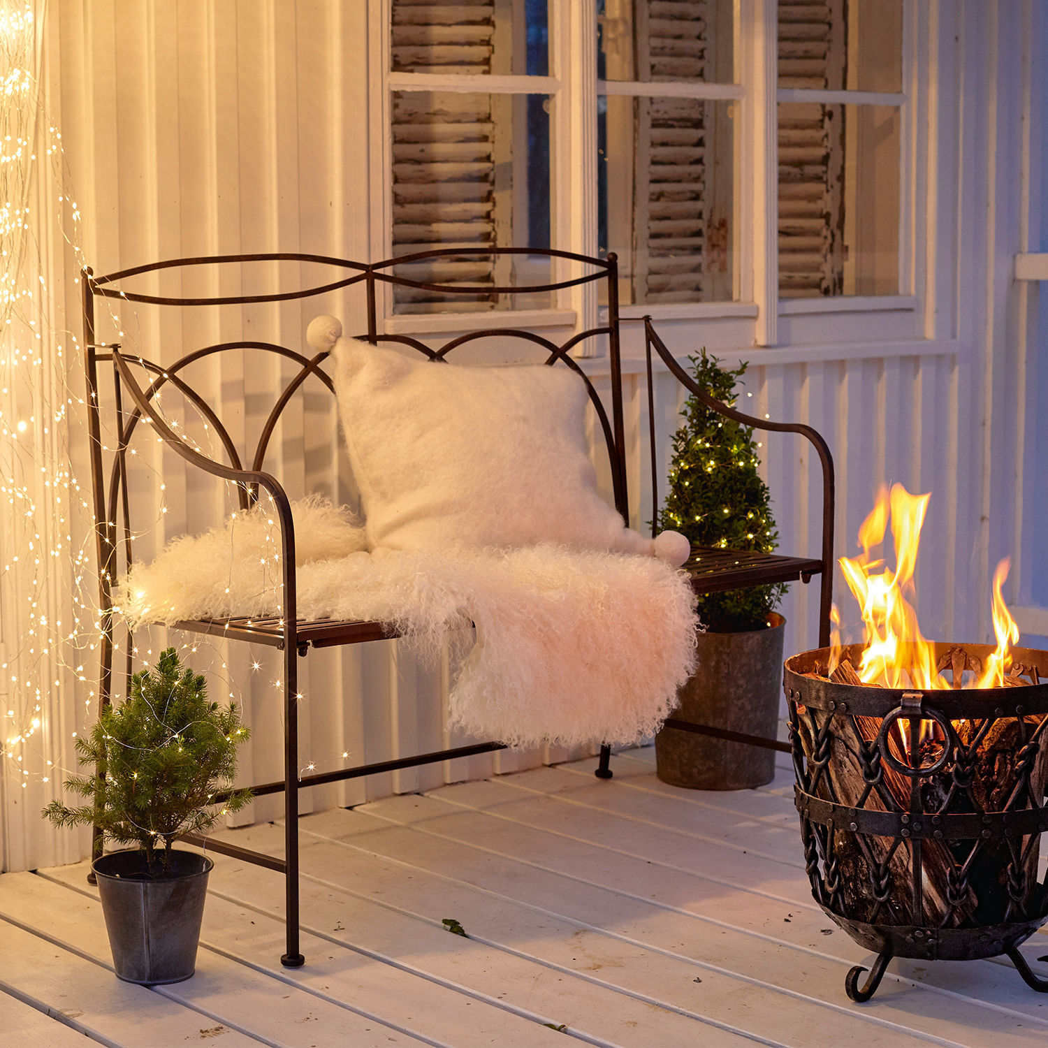 Banc en métal avec coussin blanc et couverture en fourrure près d'un brasero sur une terrasse décorée de guirlandes lumineuses