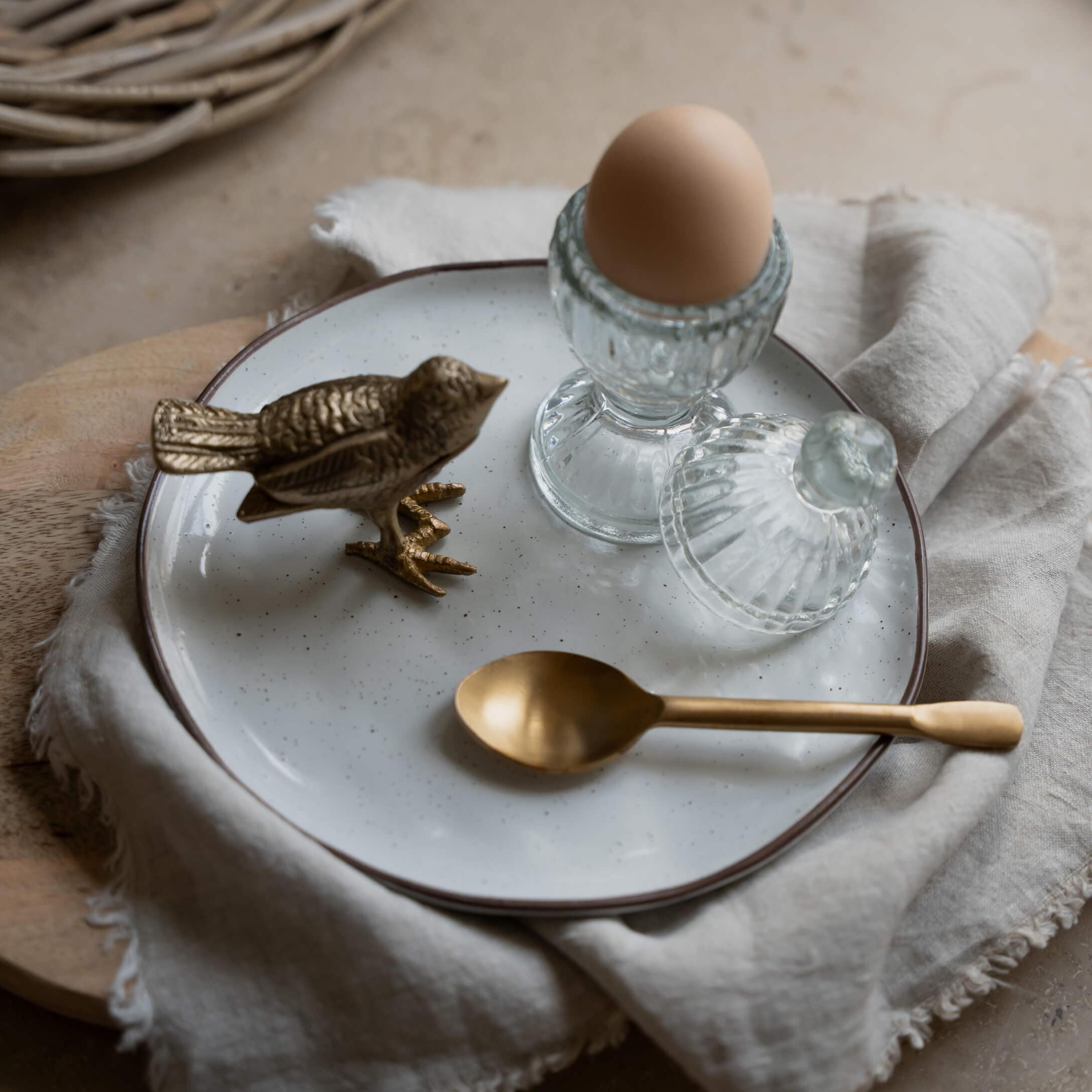 Porte-œuf en verre avec couvercle et œuf brun sur assiette blanche avec cuillère en laiton et oiseau décoratif