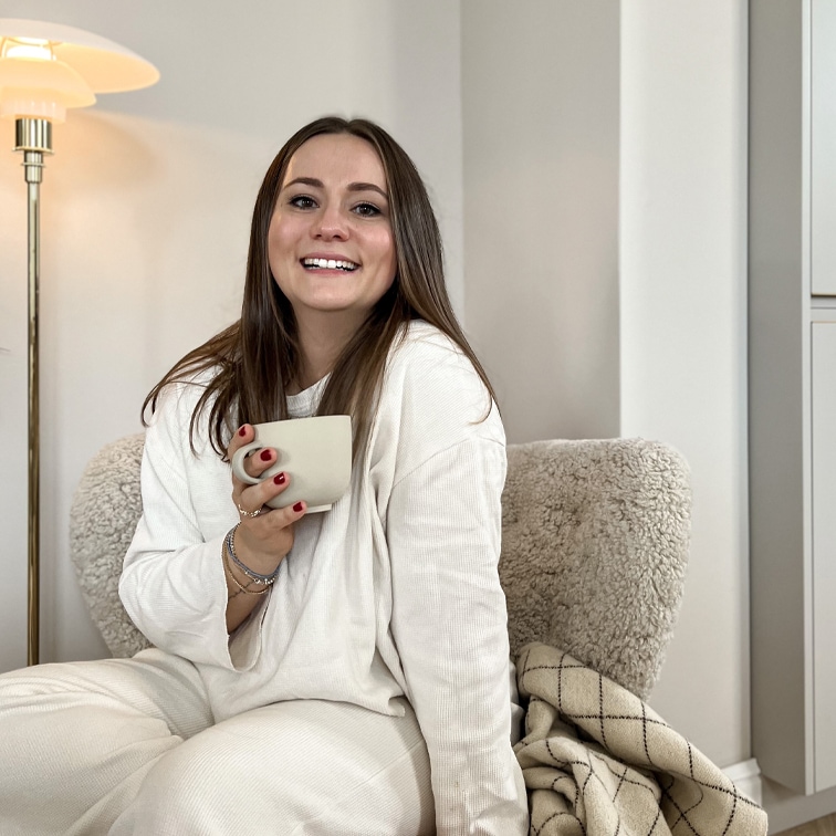 Femme souriante en tenue blanche assise sur un fauteuil doux avec une tasse dans un intérieur chaleureux