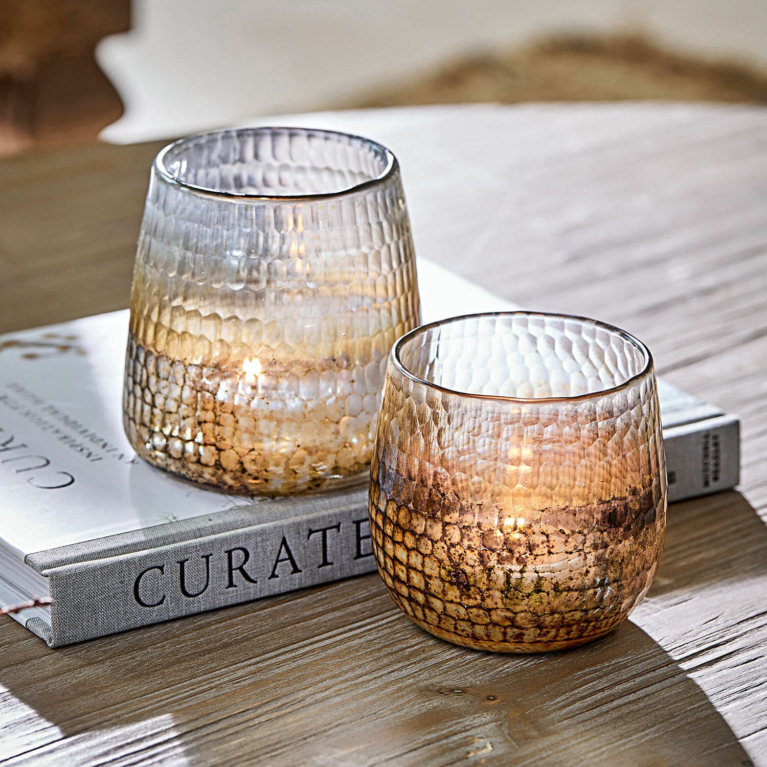 Lot de 2 photophores Agnús Deux photophores en verre texturé aux tons bruns posés sur une table en bois avec un livre