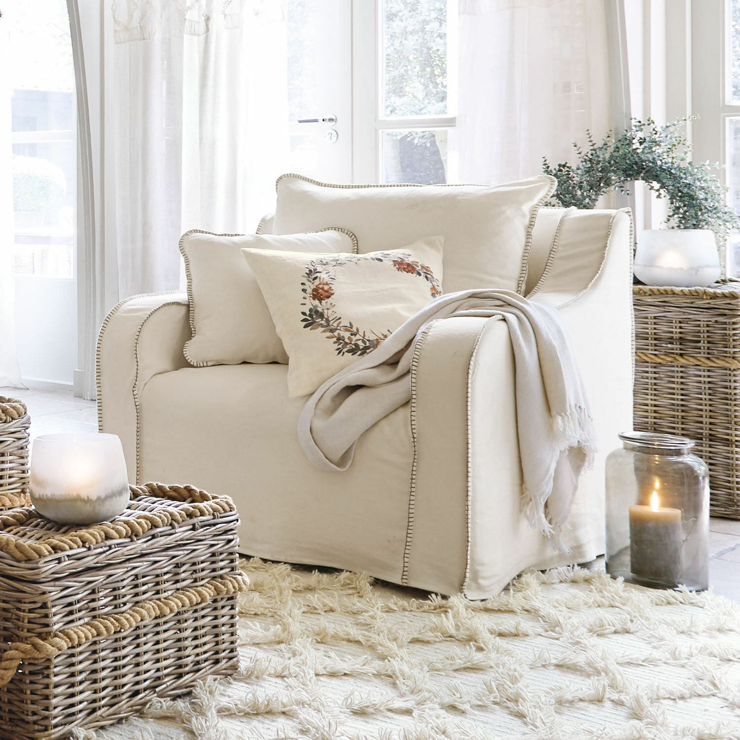 Fauteuil Ulima Fauteuil beige en tissu avec coussins et plaid dans un salon clair avec paniers en osier et bougies