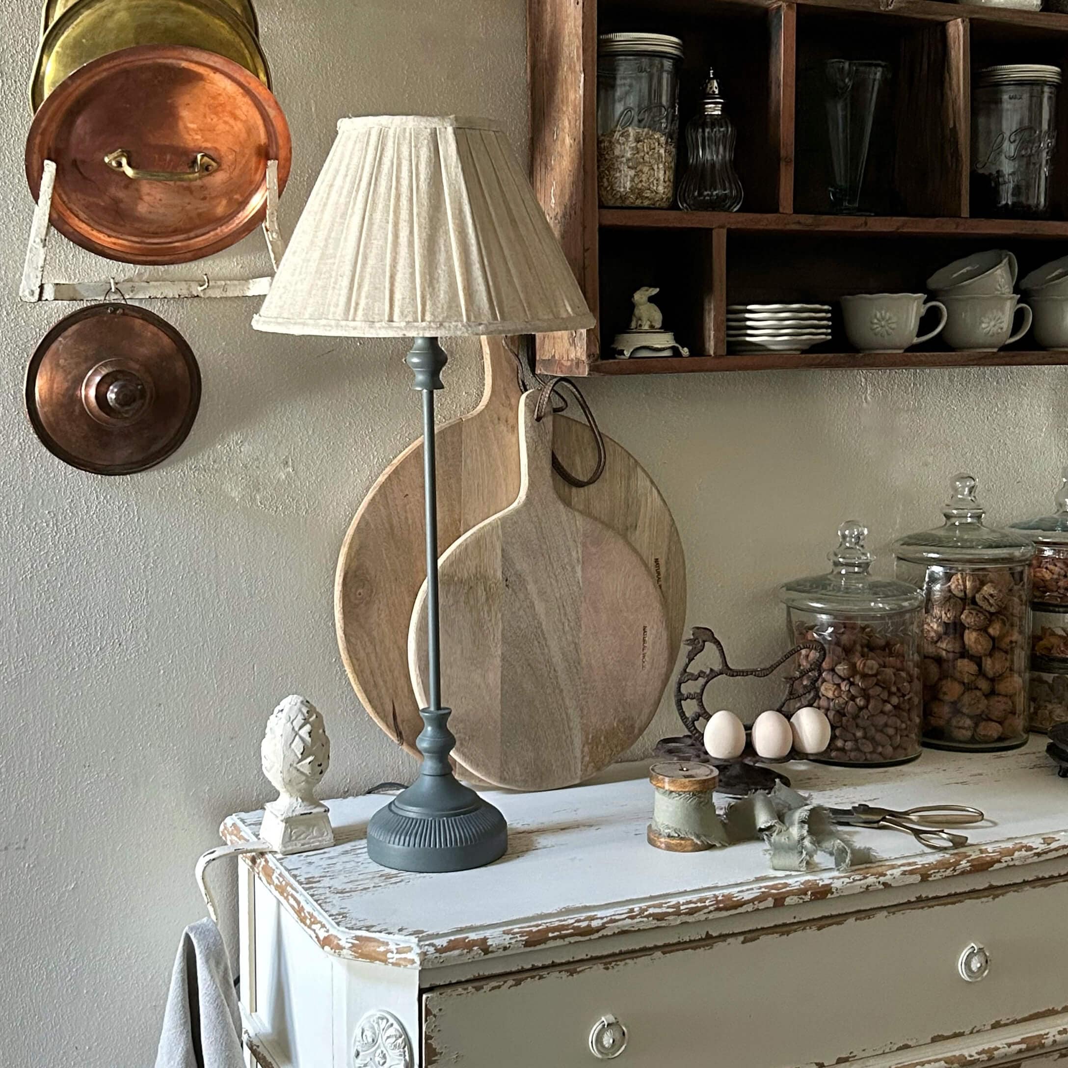 Lampe de table avec base grise et abat-jour crème sur commode blanche vieillie dans une cuisine rustique
