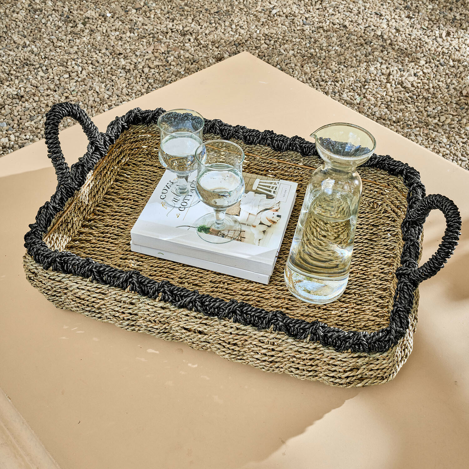 Plateau Tarevo Plateau rectangulaire en herbe de mer tressée avec bordure noire et poignées, posé avec carafe et verres sur table