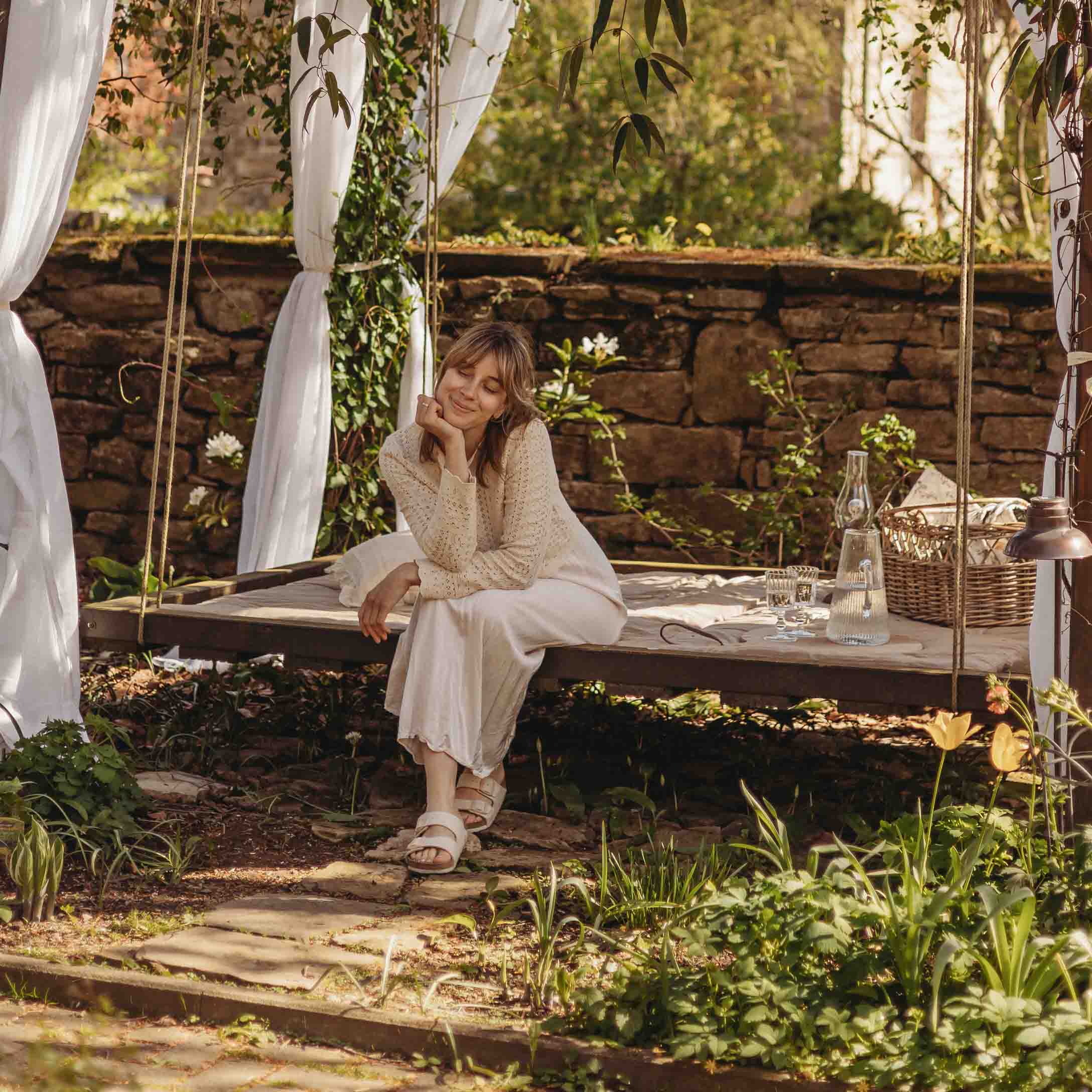 Femme en pull beige assise sur une balançoire de jardin en bois avec rideaux blancs et décor naturel