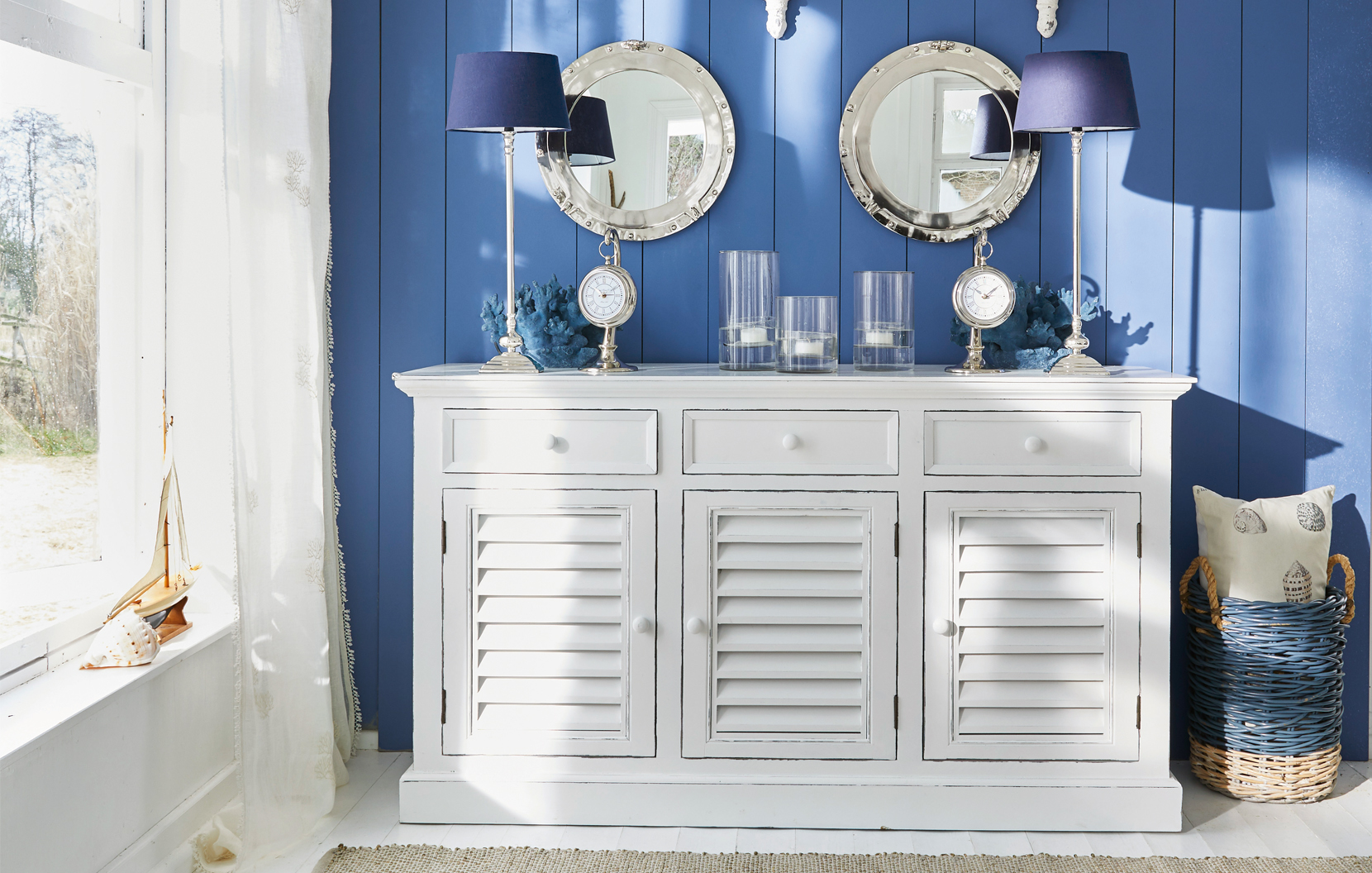 Buffet blanc en bois avec portes à persiennes sur mur bleu décoré d'objets marins