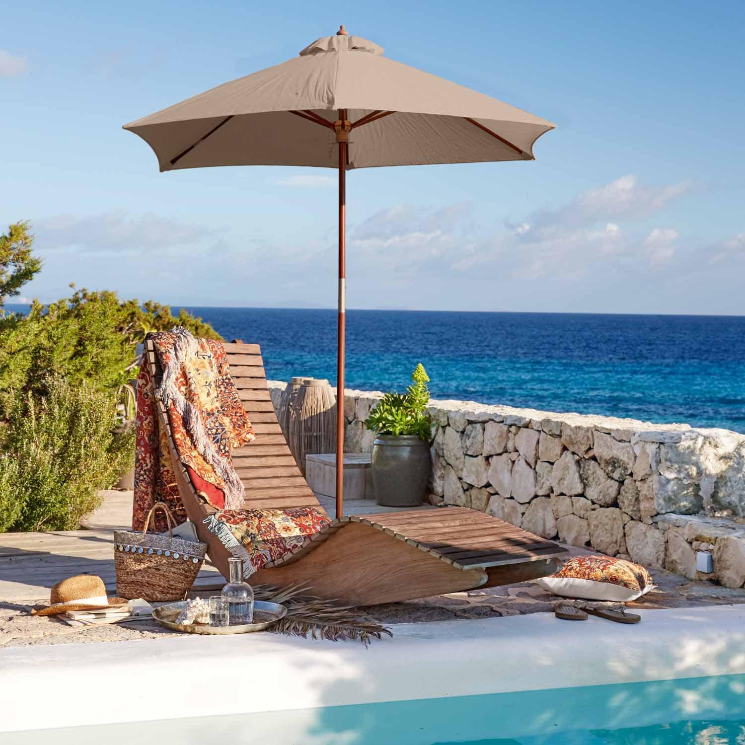 Chaise longue en bois avec parasol beige et plaid coloré au bord de la piscine face à la mer