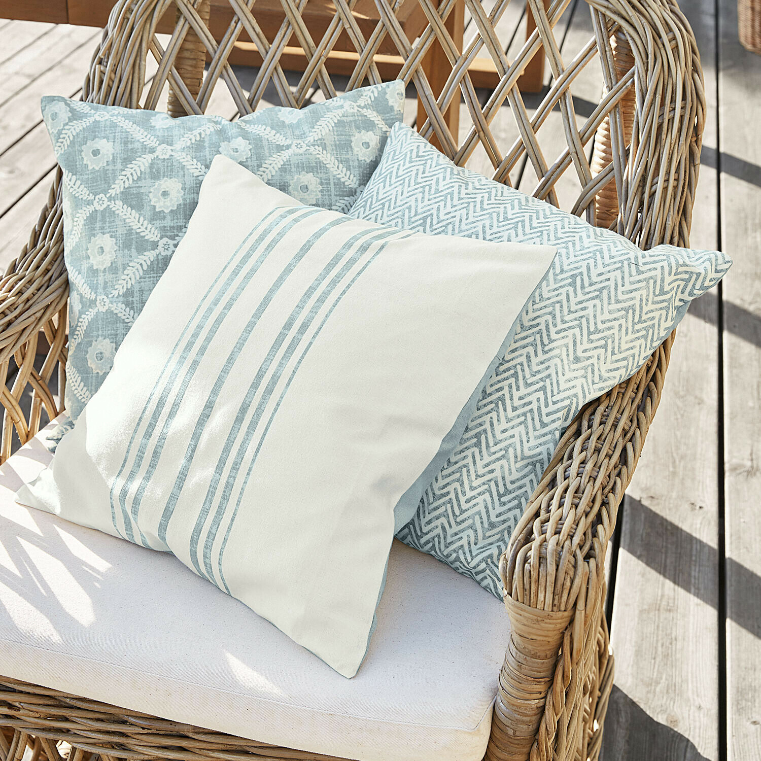Lot de 3 housses de coussins Sigruna Trois coussins décoratifs bleu clair et blanc sur un fauteuil en osier sur une terrasse en bois