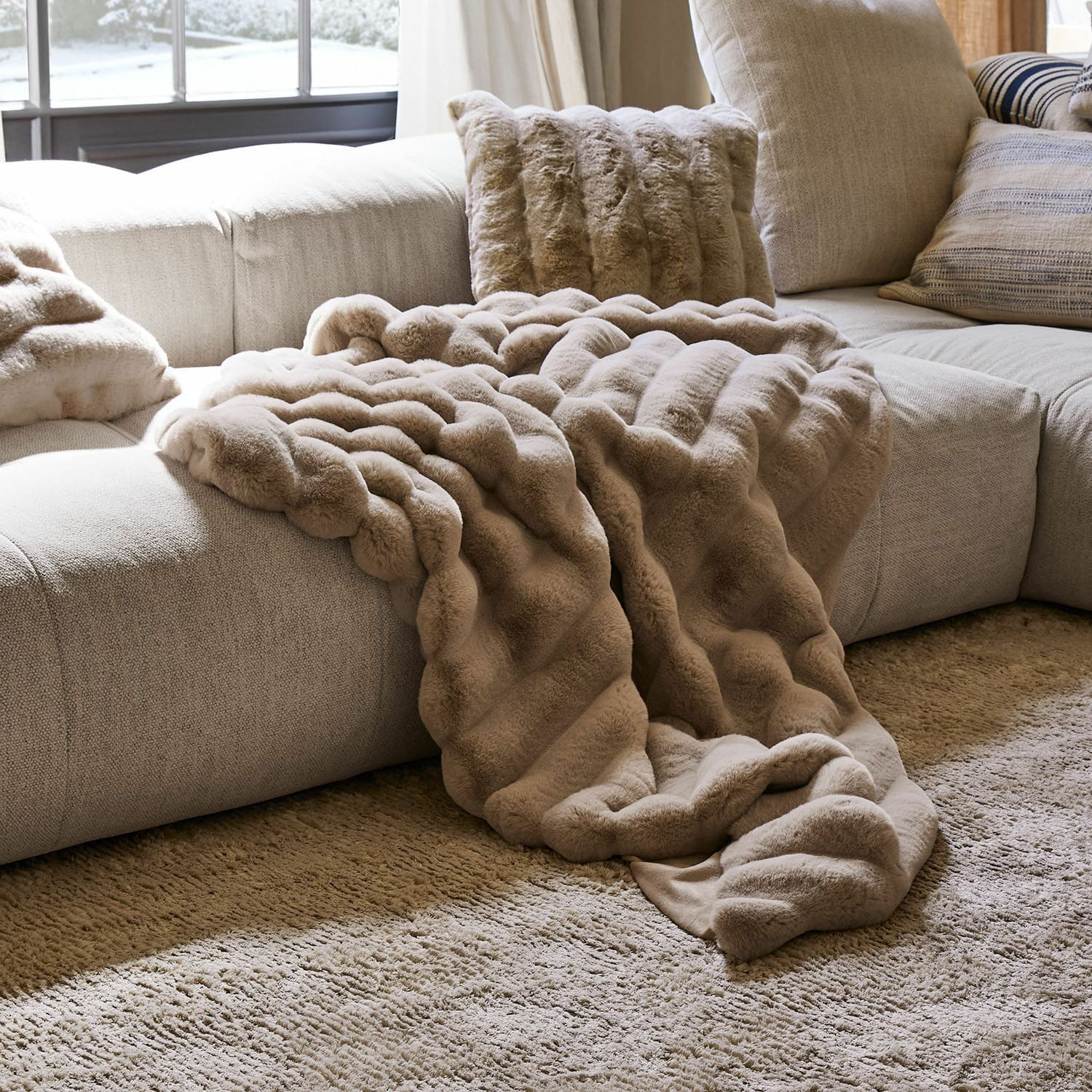 Plaid Caelira Plaid et coussin en fausse fourrure beige sur un canapé gris clair dans un salon chaleureux