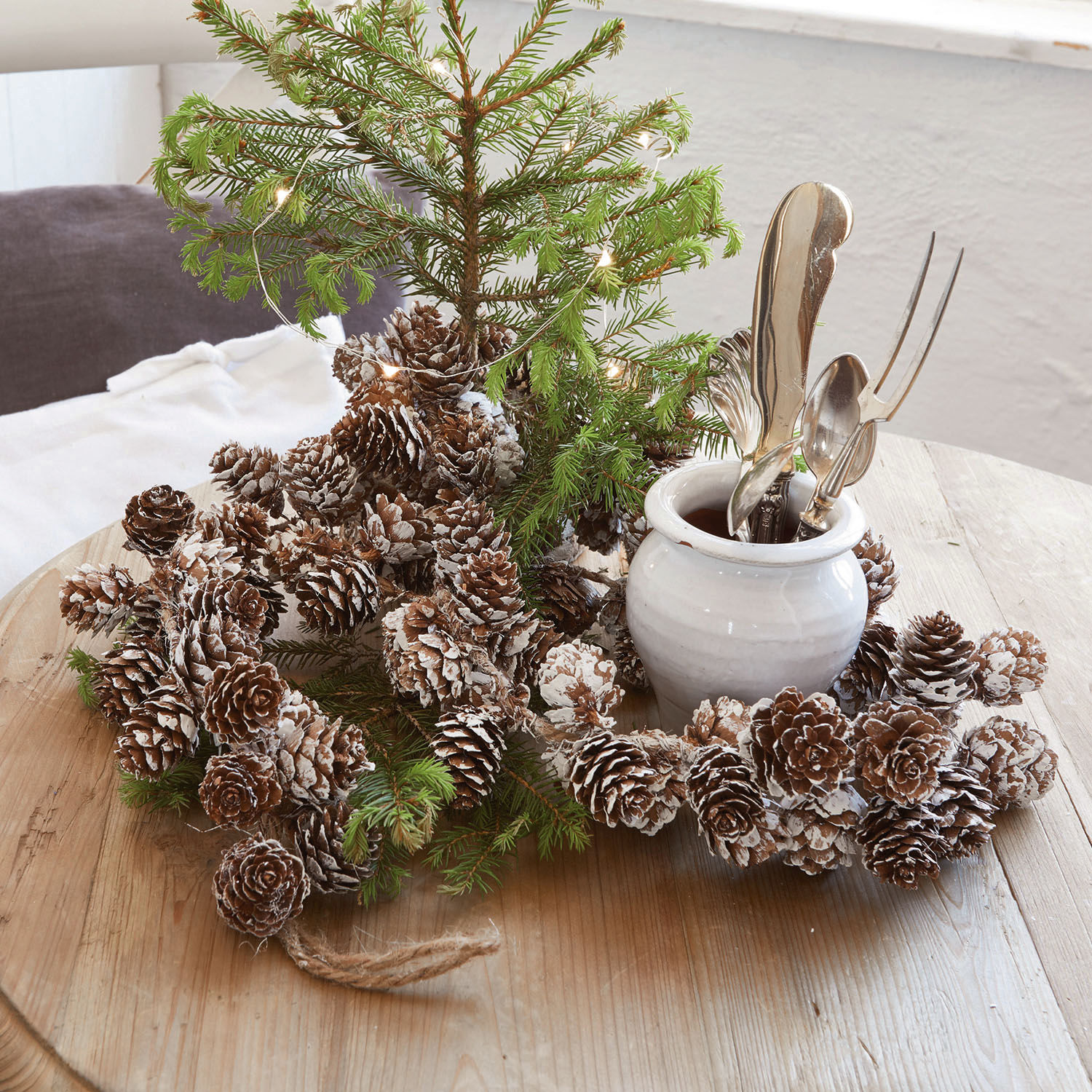 Guirlande de pommes de pin Jacci Couronne décorative en pommes de pin enneigées sur table en bois avec branches vertes et pot blanc