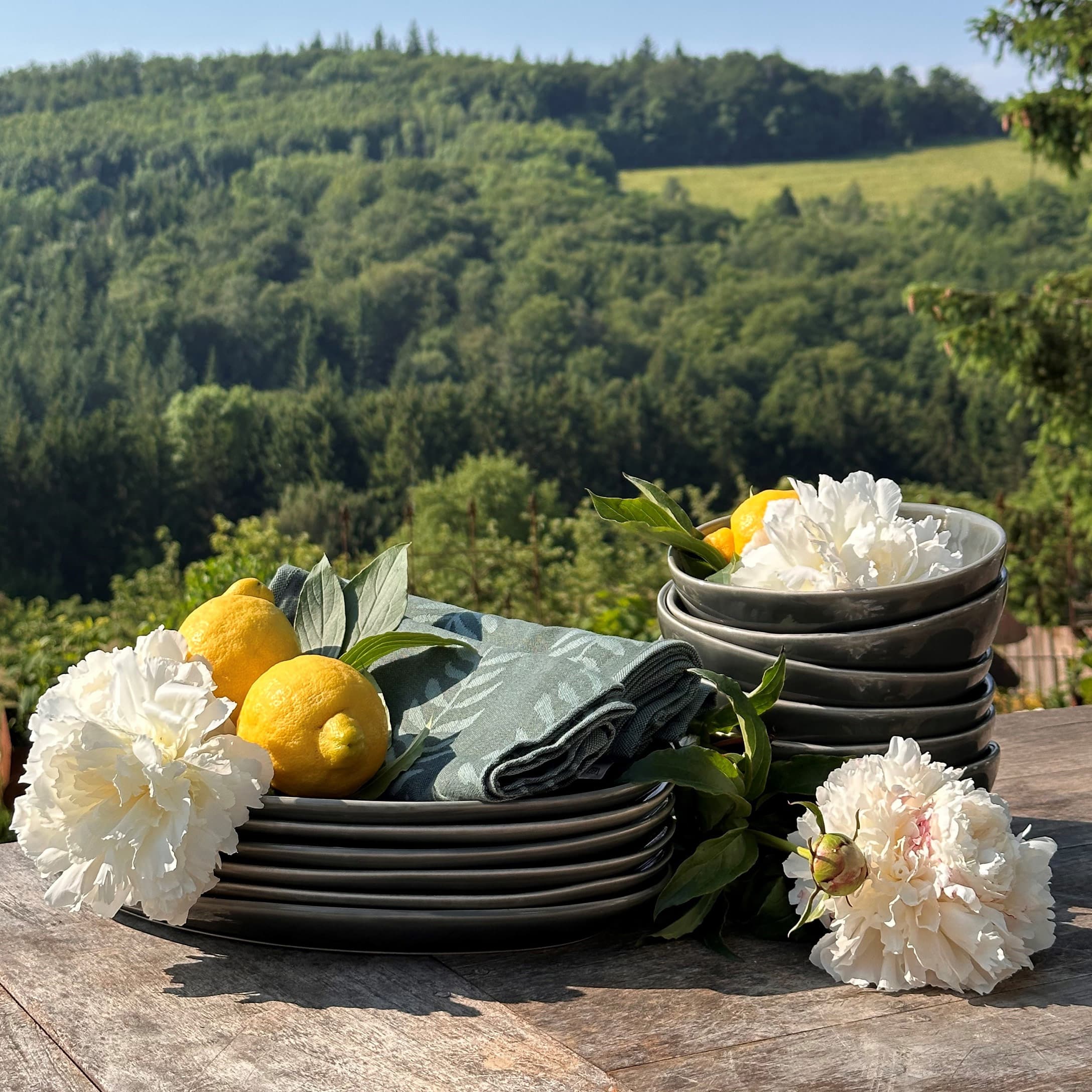 Ensemble d'assiettes et bols en céramique grise avec fleurs blanches et citrons sur table en bois devant forêt verte