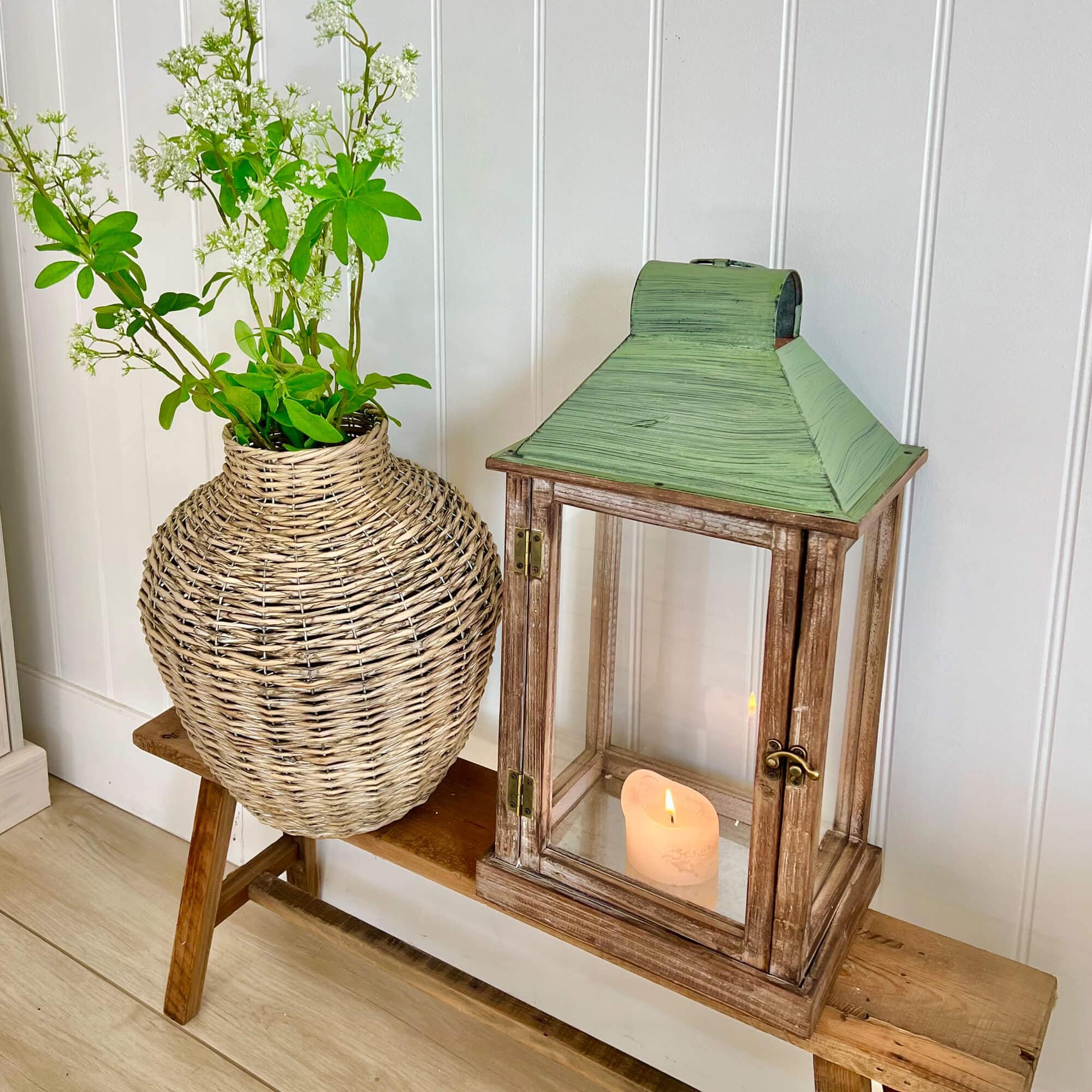 Lanterne en bois rustique avec toit en métal vert et vase en osier avec fleurs blanches sur banc en bois