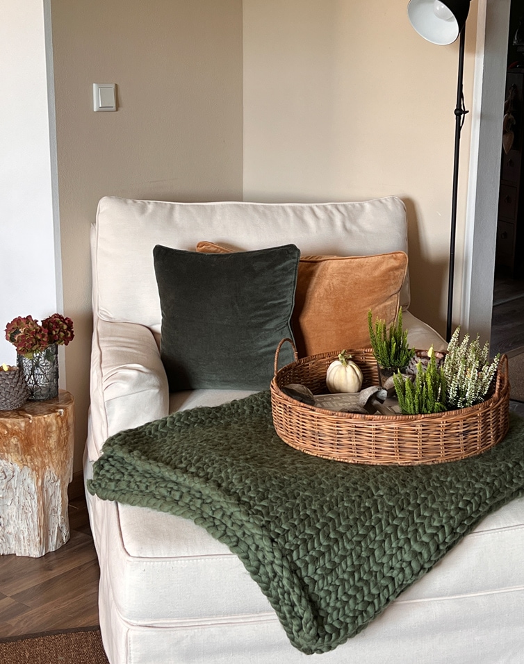 Fauteuil beige avec coussins en velours vert et orange, couverture tricotée verte et panier en osier avec décorations