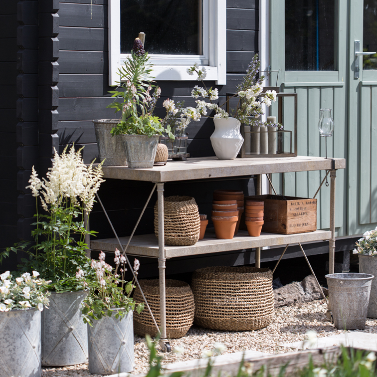 Table de jardin en métal avec pots, paniers en osier et fleurs devant un mur noir et une porte verte