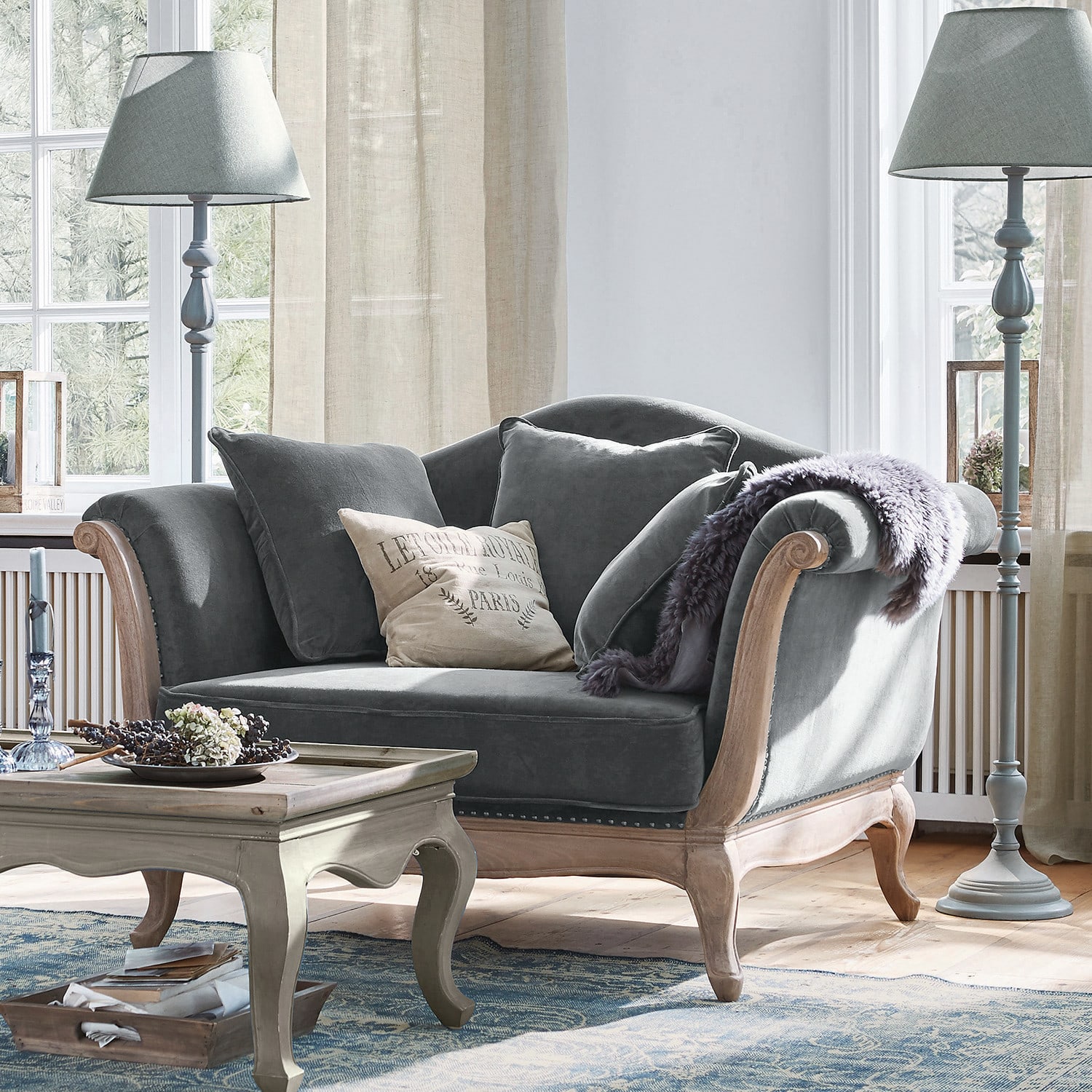 Fauteuil Sumerset Canapé gris en velours avec cadre en bois et coussins dans un salon lumineux avec lampes sur pied