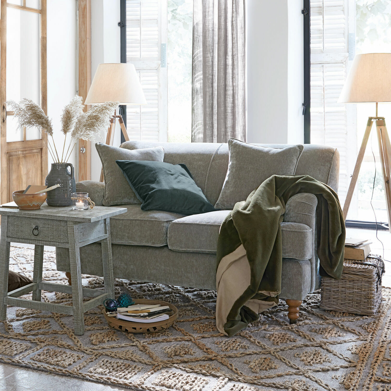 Canapé gris avec coussins doux et plaid vert dans un salon lumineux avec lampes en bois et tapis