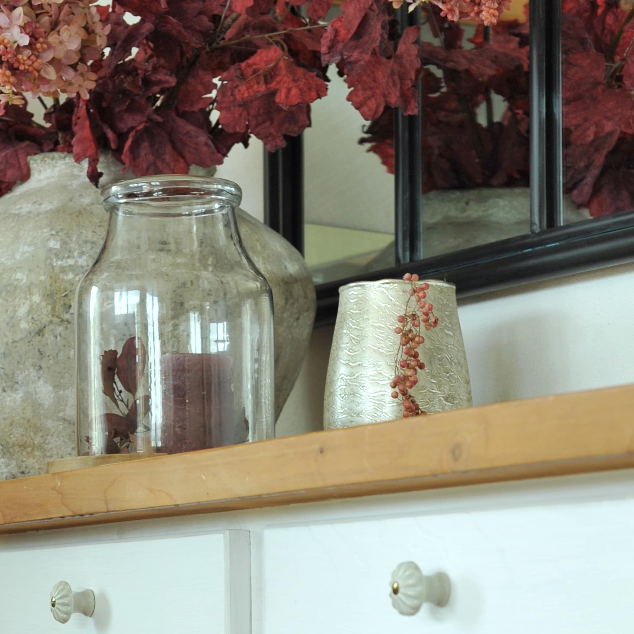 Vase en verre transparent avec bougie bordeaux sur étagère en bois décorée de feuilles rouges