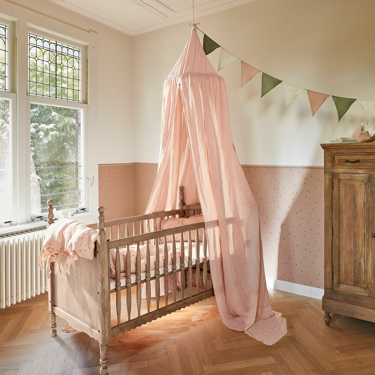 Lit bébé Merveillon Lit bébé en bois avec ciel de lit rose et literie assortie dans une chambre d'enfant pastel