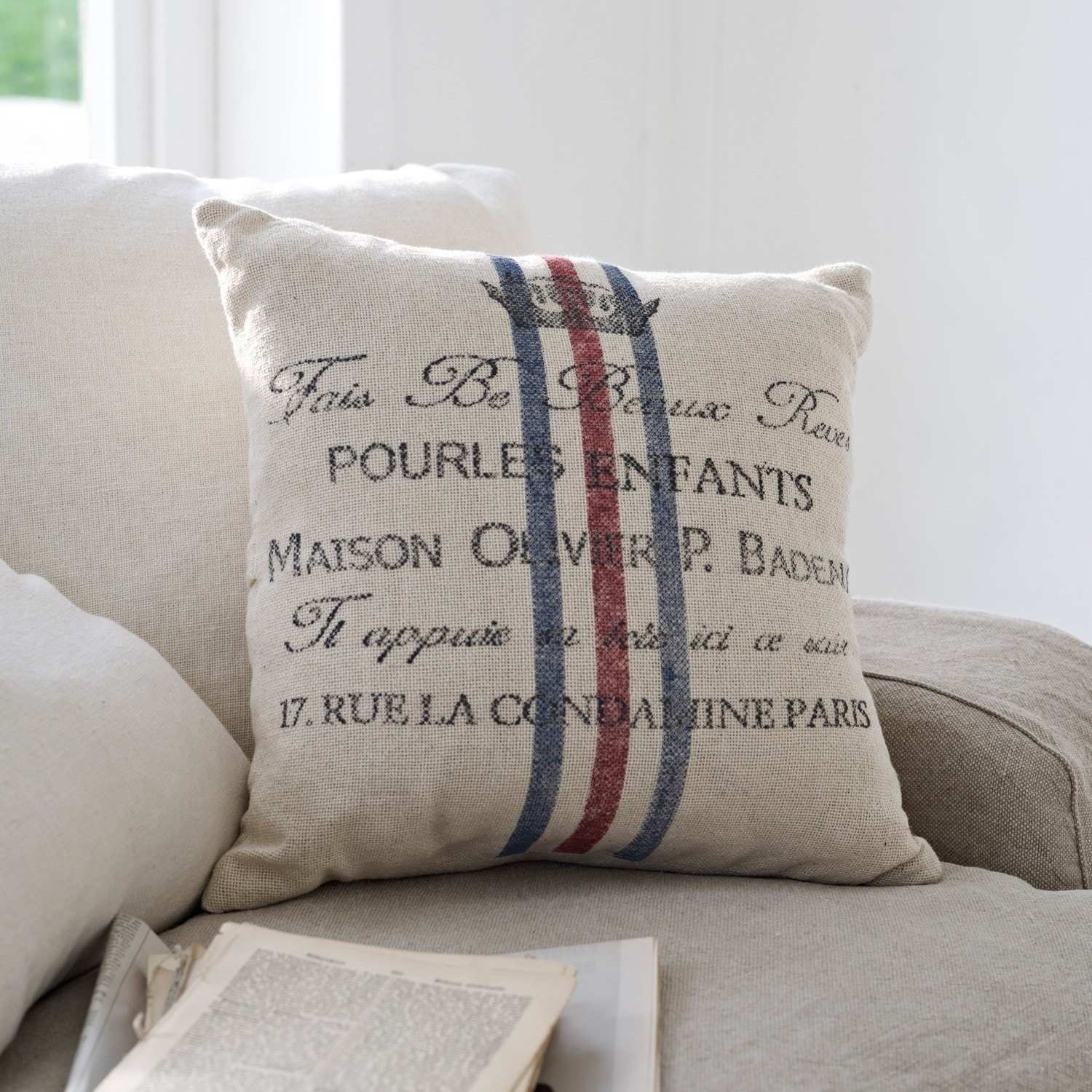 Coussin Pourl Coussin beige en tissu avec impression et bandes rouges et bleues sur canapé clair dans un salon