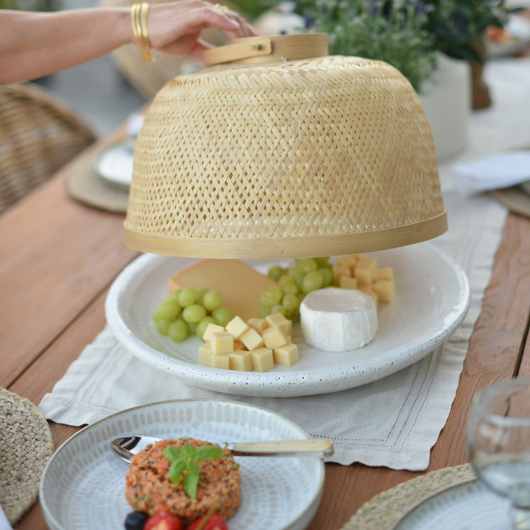 Cloche en bambou naturel protégeant fromage et raisins verts sur assiette blanche posée sur table en bois