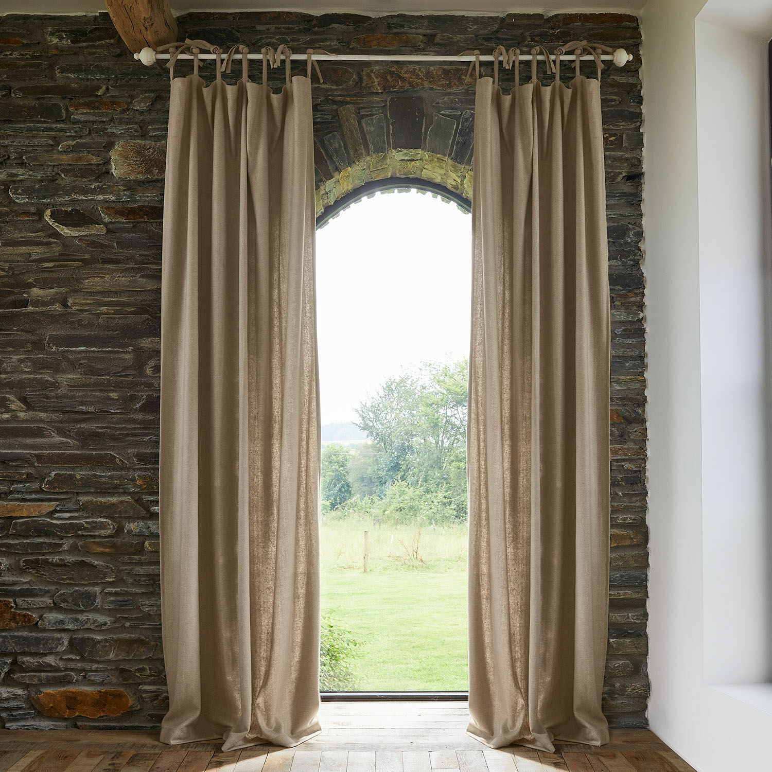 Rideau Seleneia Rideaux beiges en tissu naturel suspendus sur une tringle dans un intérieur en pierre avec vue sur le jardin